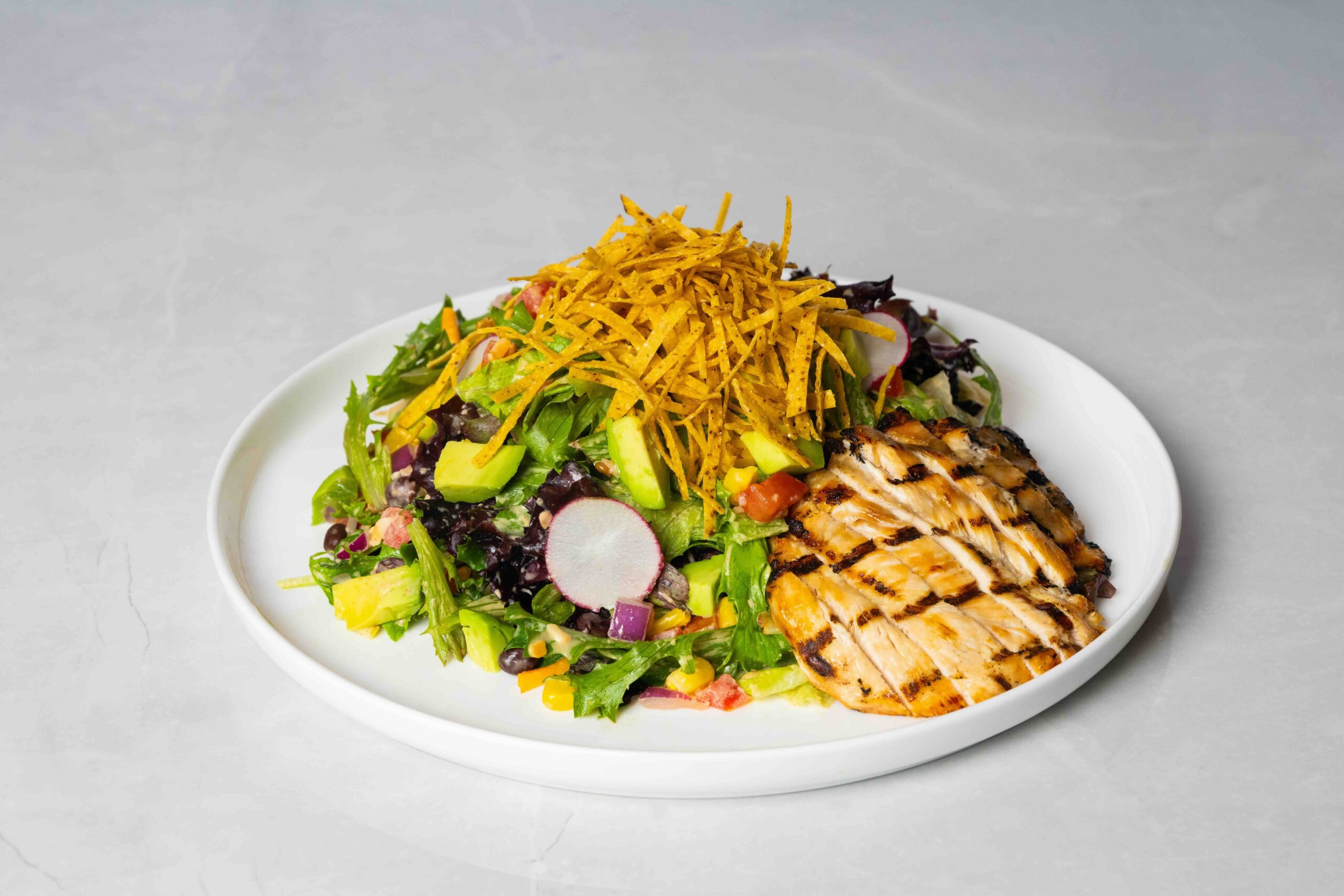 CRAFT Beer Market A plate of grilled chicken breast slices served with a mixed salad, avocado, radishes, black beans, and crispy tortilla strips—perfect for casual dining or pairing with your favorite craft beer in our beer garden.