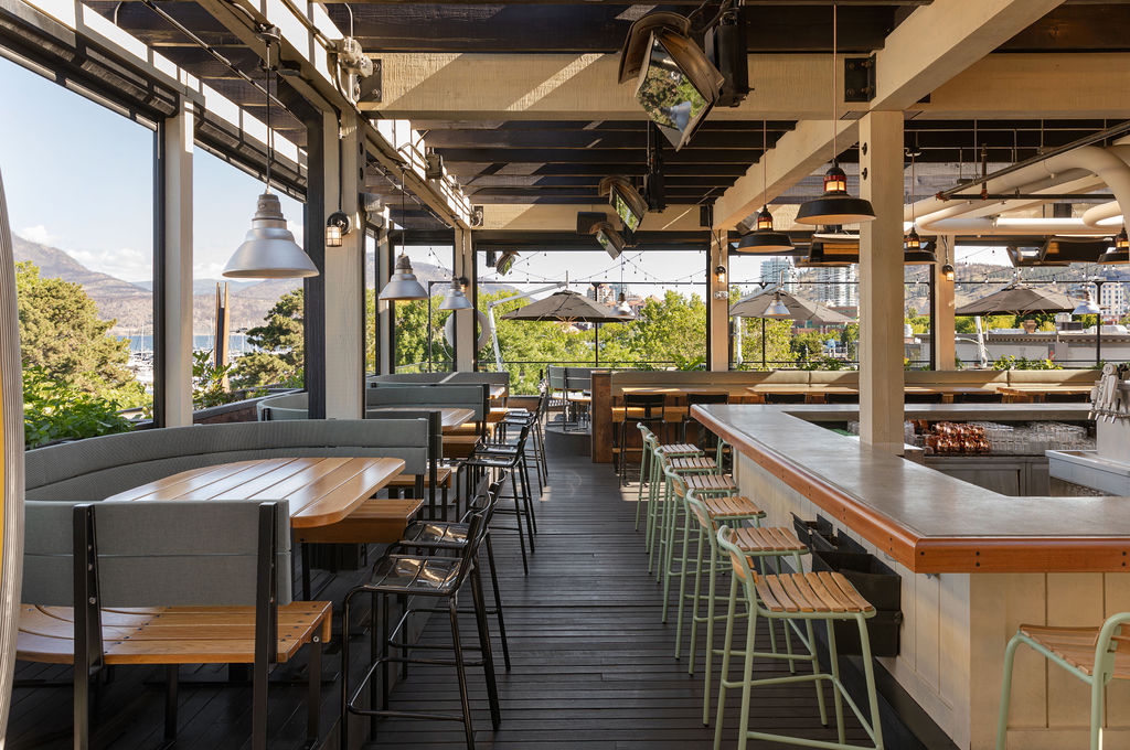 CRAFT Beer Market Outdoor restaurant patio with wooden tables, bar seating, and metal chairs, overlooking trees and city buildings in the background on a sunny day.