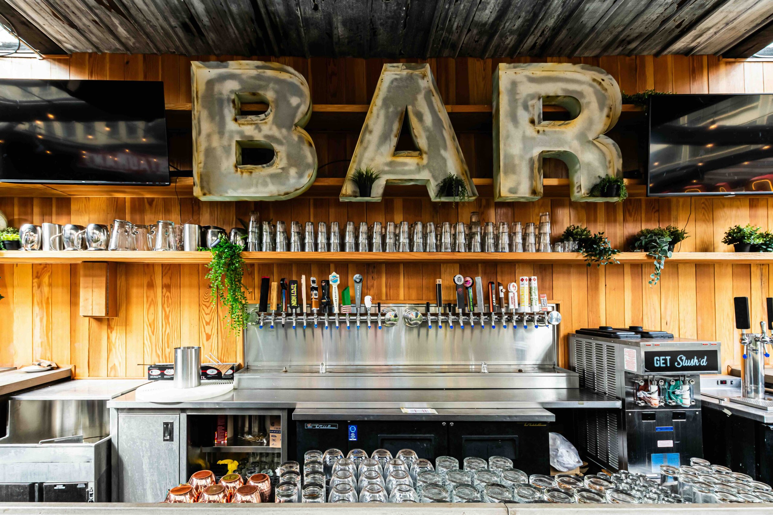 CRAFT Beer Market A bar with large illuminated "BAR" letters above beer taps, glassware, and plants on wooden shelves, with two TVs visible on either side.