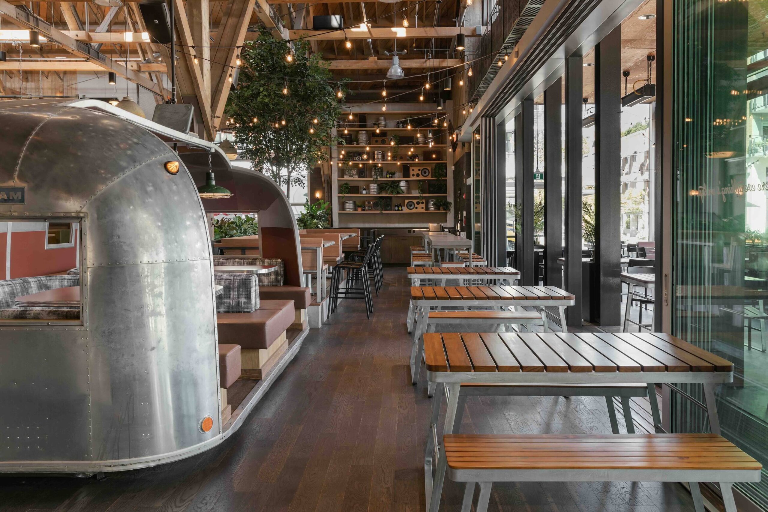 CRAFT Beer Market Modern restaurant interior with wood tables, metal benches, string lights, and a vintage silver Airstream trailer dining booth. Large windows line one side, creating perfect social vibes for casual dining or enjoying a craft beer.
