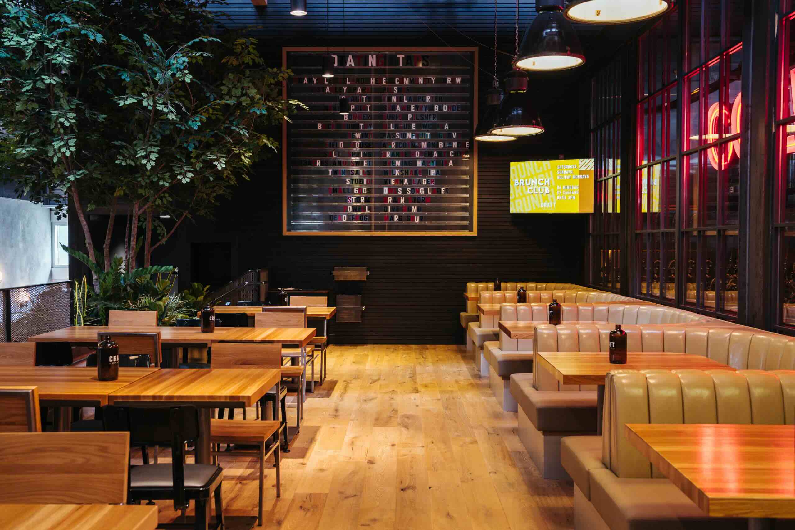 CRAFT Beer Market Empty restaurant interior with wooden tables, tan booths, a large menu board on the back wall, and plants near the seating area.