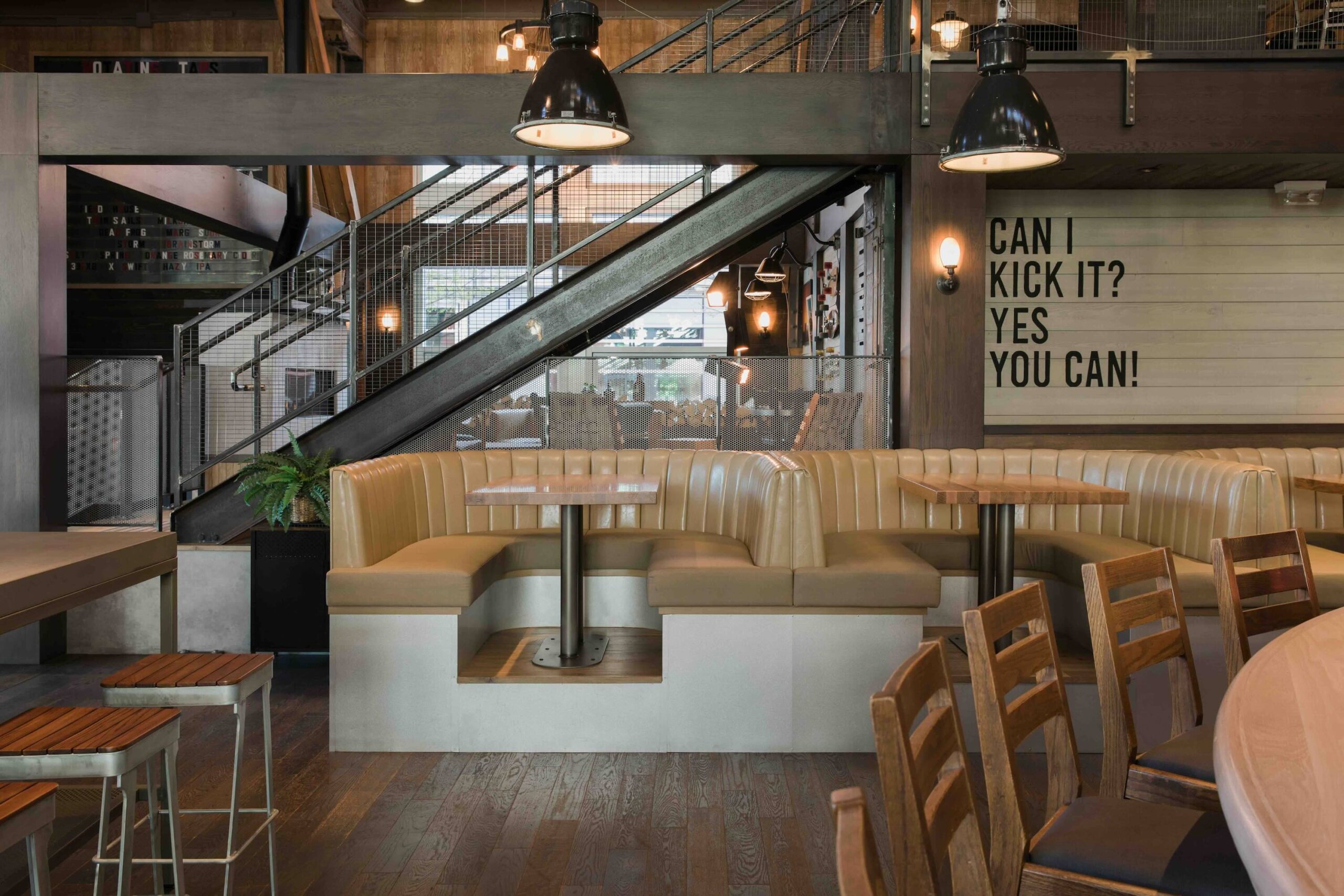 CRAFT Beer Market Restaurant interior with beige booth seating, wooden chairs, industrial lighting, and a wall sign that reads "CAN I KICK IT? YES YOU CAN!" next to a staircase—perfect for casual dining and social vibes.