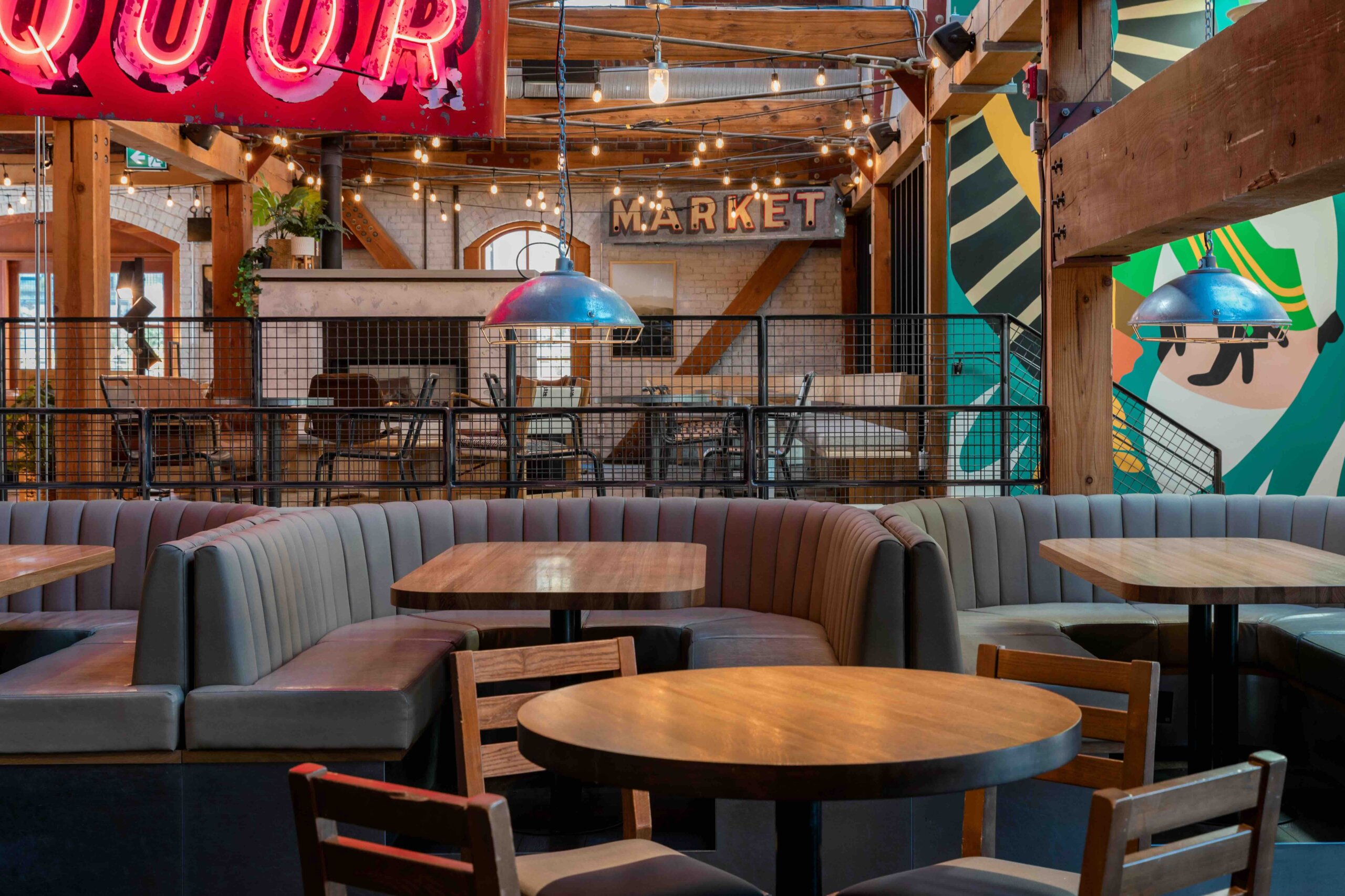 CRAFT Beer Market Modern indoor dining area with wooden tables, gray upholstered booths, neon signage, hanging lights, and colorful wall art.