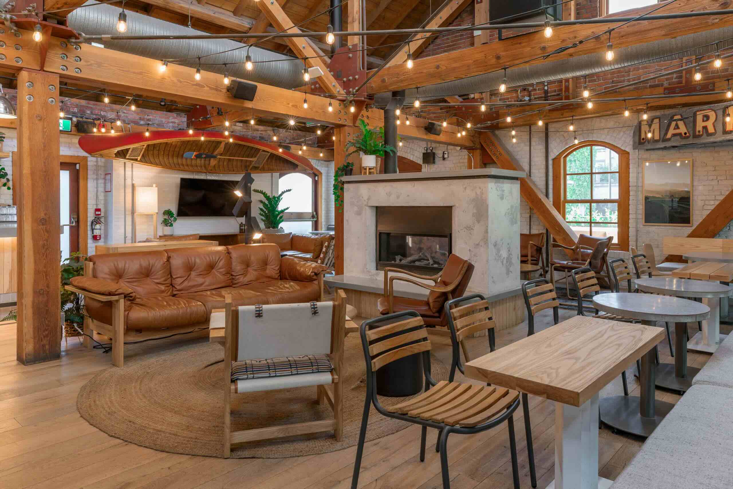 CRAFT Beer Market Spacious lounge with wooden beams, leather couches, modern chairs, string lights, and a central fireplace, featuring large windows and indoor plants.