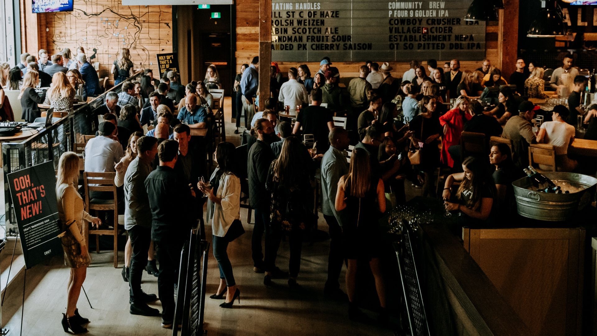CRAFT Beer Market A crowded bar or restaurant with people socializing, standing and sitting at tables, with a menu board visible on the wooden wall.