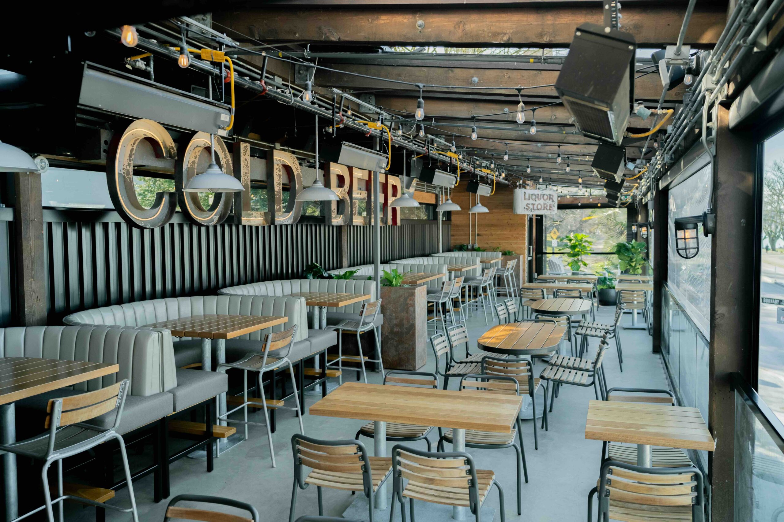 CRAFT Beer Market Outdoor bar seating area with wooden tables, chairs, cushioned booths, string lights, and large "COLD BEER" sign on the wall. Space is empty and well-lit by natural light.
