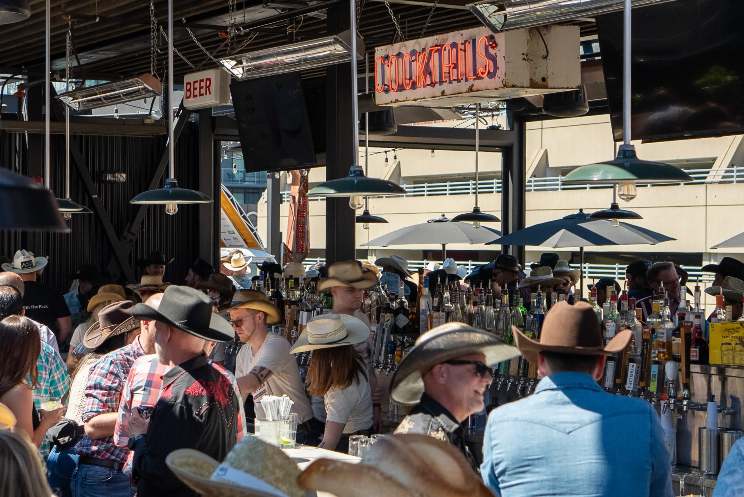 CRAFT Beer Market Crowded bar scene with people in cowboy hats socializing under covered outdoor area. Signs for beer and cocktails are visible above the bar.