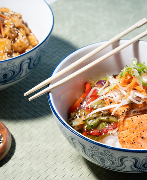 CRAFT Beer Market Two bowls of Asian-inspired dishes sit ready for casual dining; the front features rice, grilled salmon, and vegetables with chopsticks on top. Another tasty bowl is partially visible—perfect fare for enjoying in a beer garden or with craft cocktails.