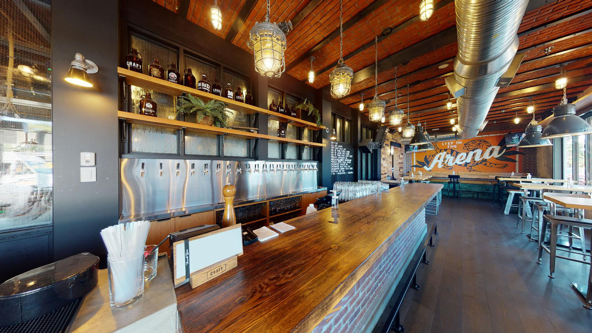 CRAFT Beer Market A modern bar with a long wooden counter, multiple beer taps, shelves with bottles, industrial lighting, and a mural that reads “The Arena” on a brick wall. Perfect for private events or group bookings.