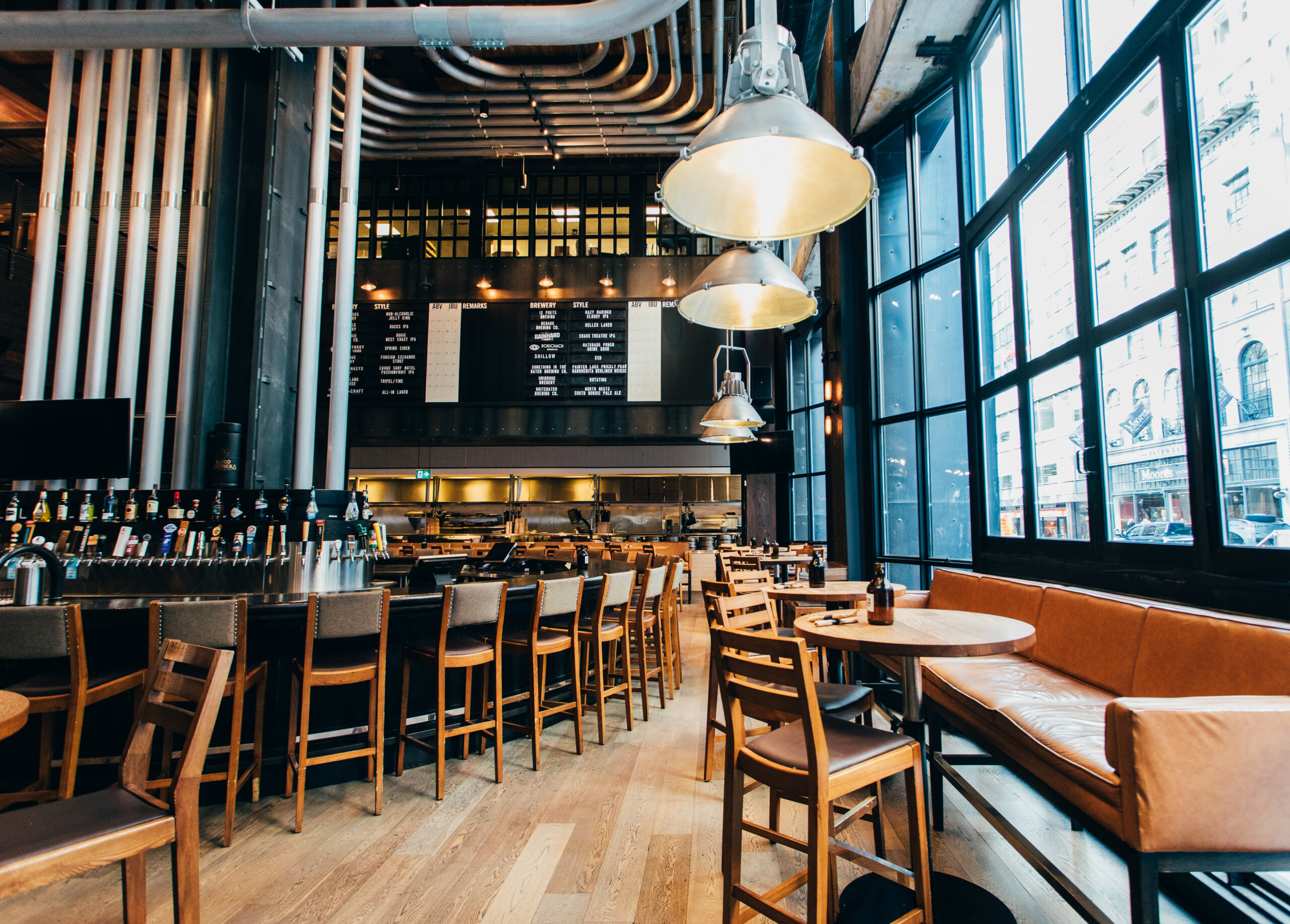 CRAFT Beer Market Modern restaurant interior with wooden tables and chairs, a long bar serving craft cocktails, industrial-style lighting, and large windows letting in natural light.
