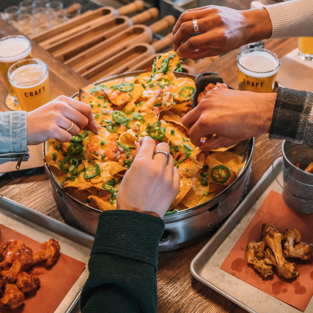 CRAFT Beer Market Four people reach for nachos topped with cheese and jalapeños, surrounded by craft beers and trays of chicken wings on a wooden table—perfect for group bookings.