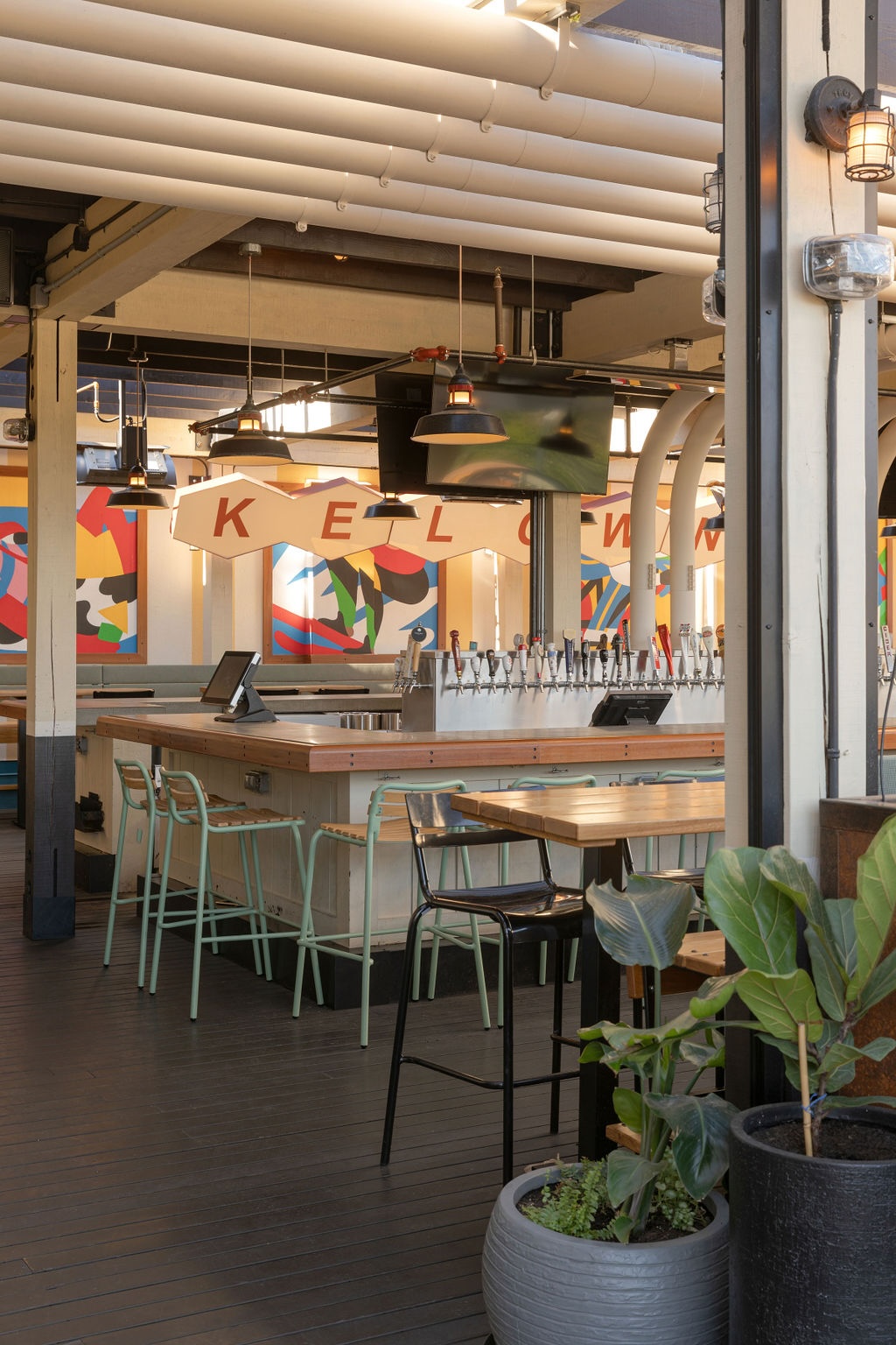 CRAFT Beer Market Modern bar area with light wood counters, high metal stools, multiple taps, colorful abstract wall art, and potted plants in a well-lit space.