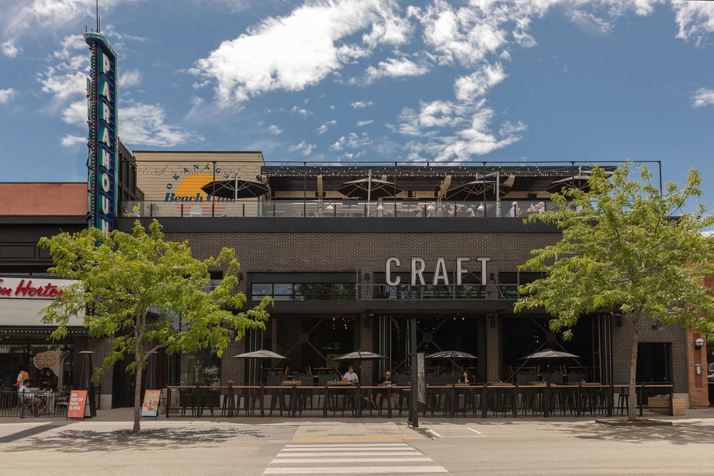 CRAFT Beer Market Street view of a modern restaurant called Craft with outdoor seating under large umbrellas, trees lining the sidewalk, and a clear blue sky overhead.