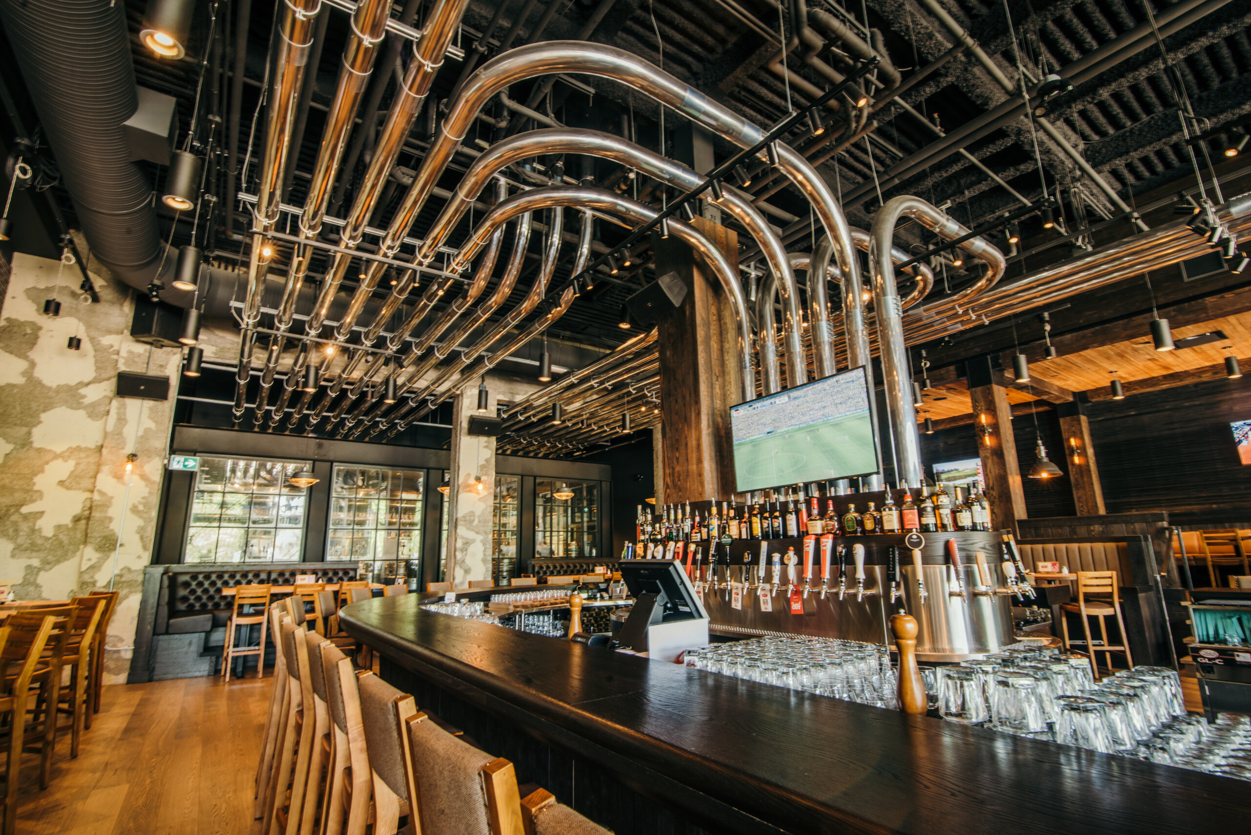 CRAFT Beer Market Modern bar interior with a long counter, wooden chairs, glassware, numerous rotational taps for beer, exposed pipes on the ceiling, and a television above the bar—perfect for enjoying social vibes.