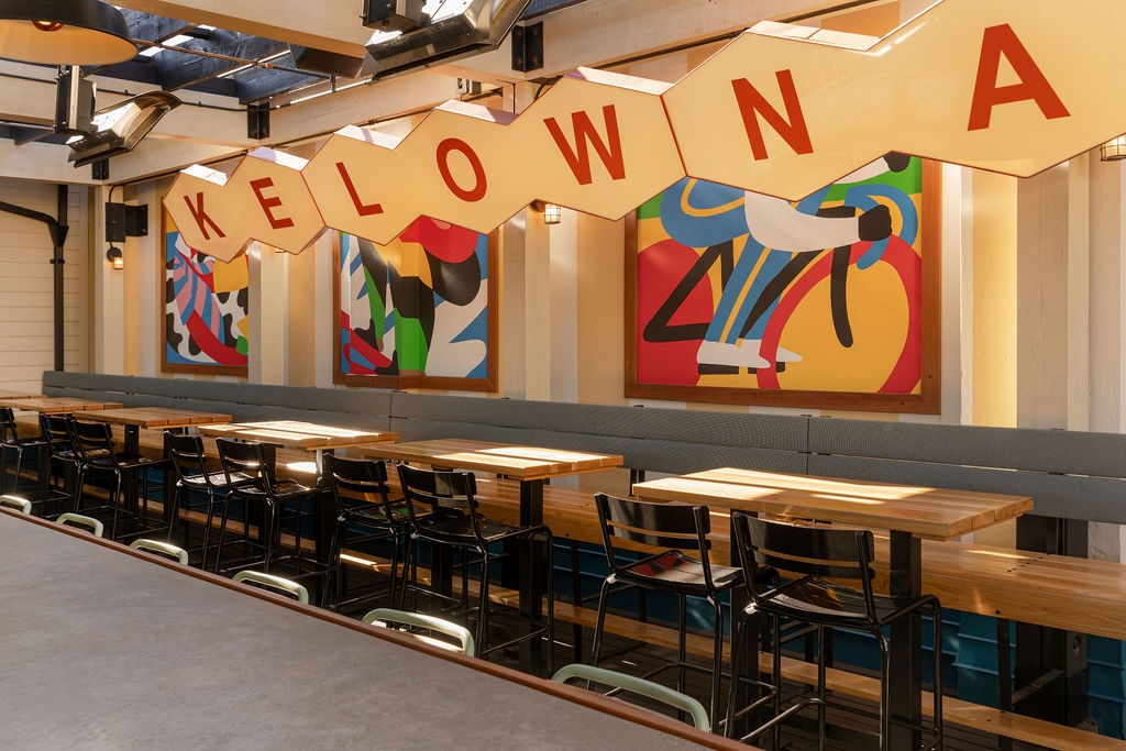 CRAFT Beer Market A row of wooden tables and black chairs sit beneath a large Kelowna sign and colorful abstract murals in a modern indoor dining area.