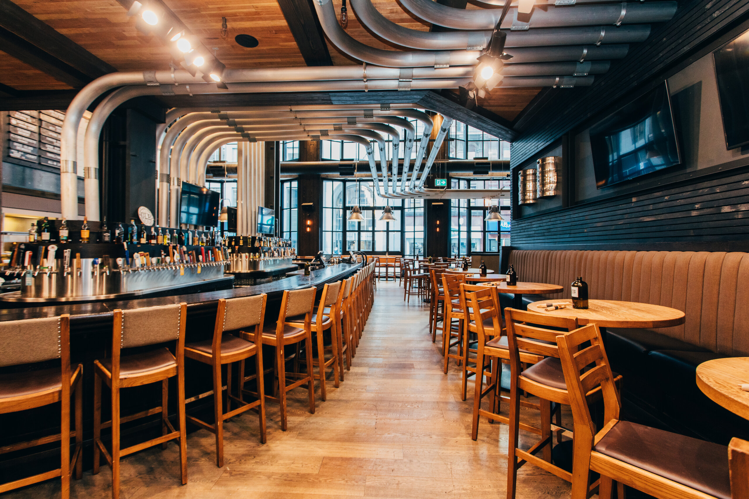 CRAFT Beer Market Modern bar interior with wooden floors, high-backed chairs at a long bar, round tables, and exposed metal pipes. Large windows let in natural light—perfect for enjoying craft beer or hosting private events in style.
