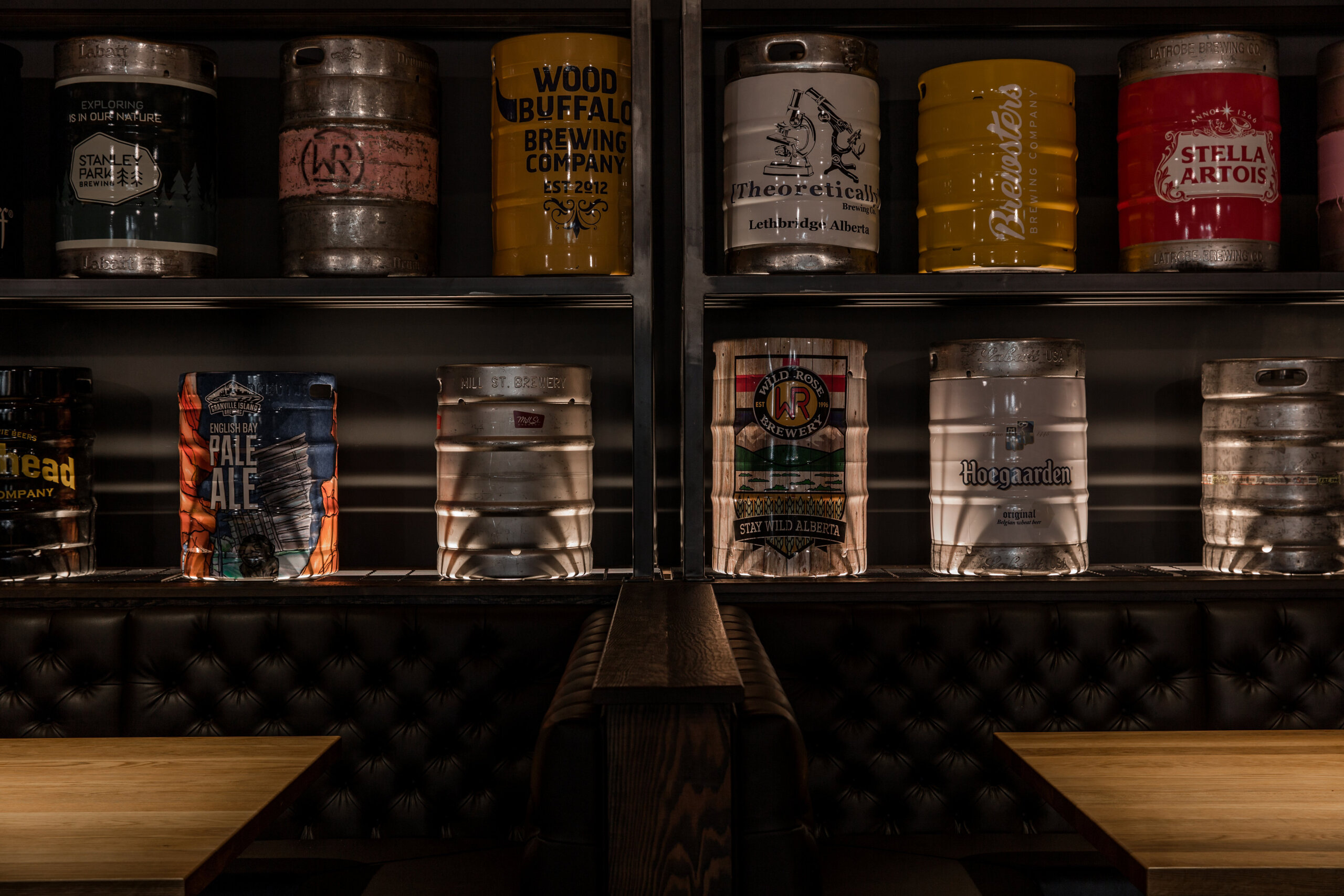 CRAFT Beer Market Rows of beer kegs and barrels, labeled with various brewery names, line the shelves above dark leather seating in a dimly lit bar—perfect for enjoying craft cocktails and soaking up the social vibes.