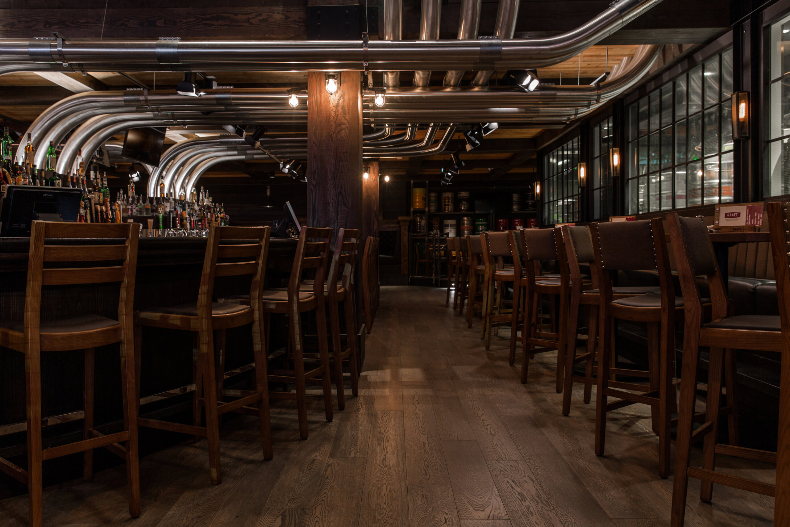 CRAFT Beer Market A modern bar interior with wooden chairs and tables, a long bar counter, rotational taps, metal pipes on the ceiling, and dim lighting.