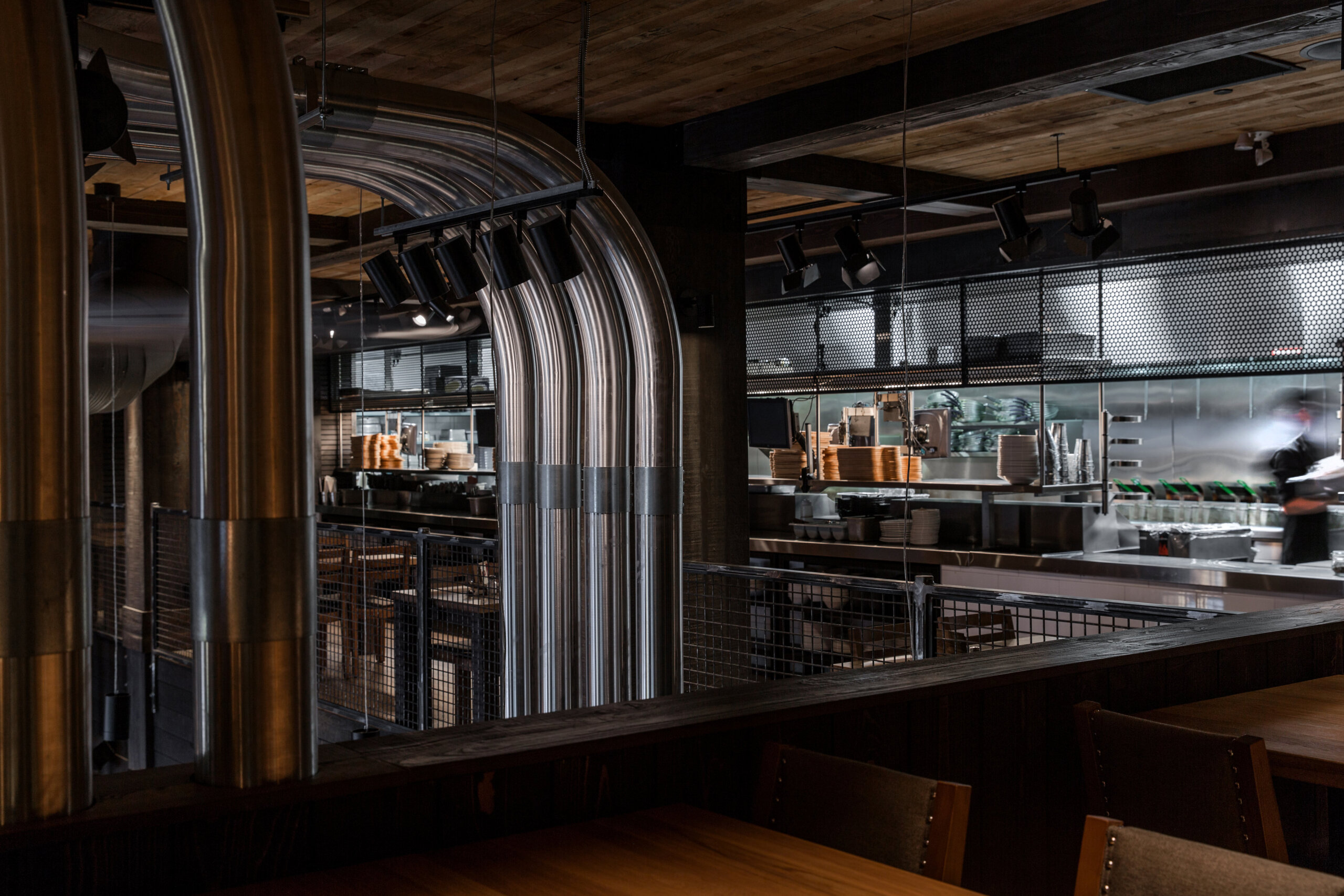 CRAFT Beer Market Modern restaurant interior with exposed metal pipes, wooden ceiling, and an open kitchen visible in the background, perfect for enjoying craft beer from rotational taps and soaking in the social vibes.