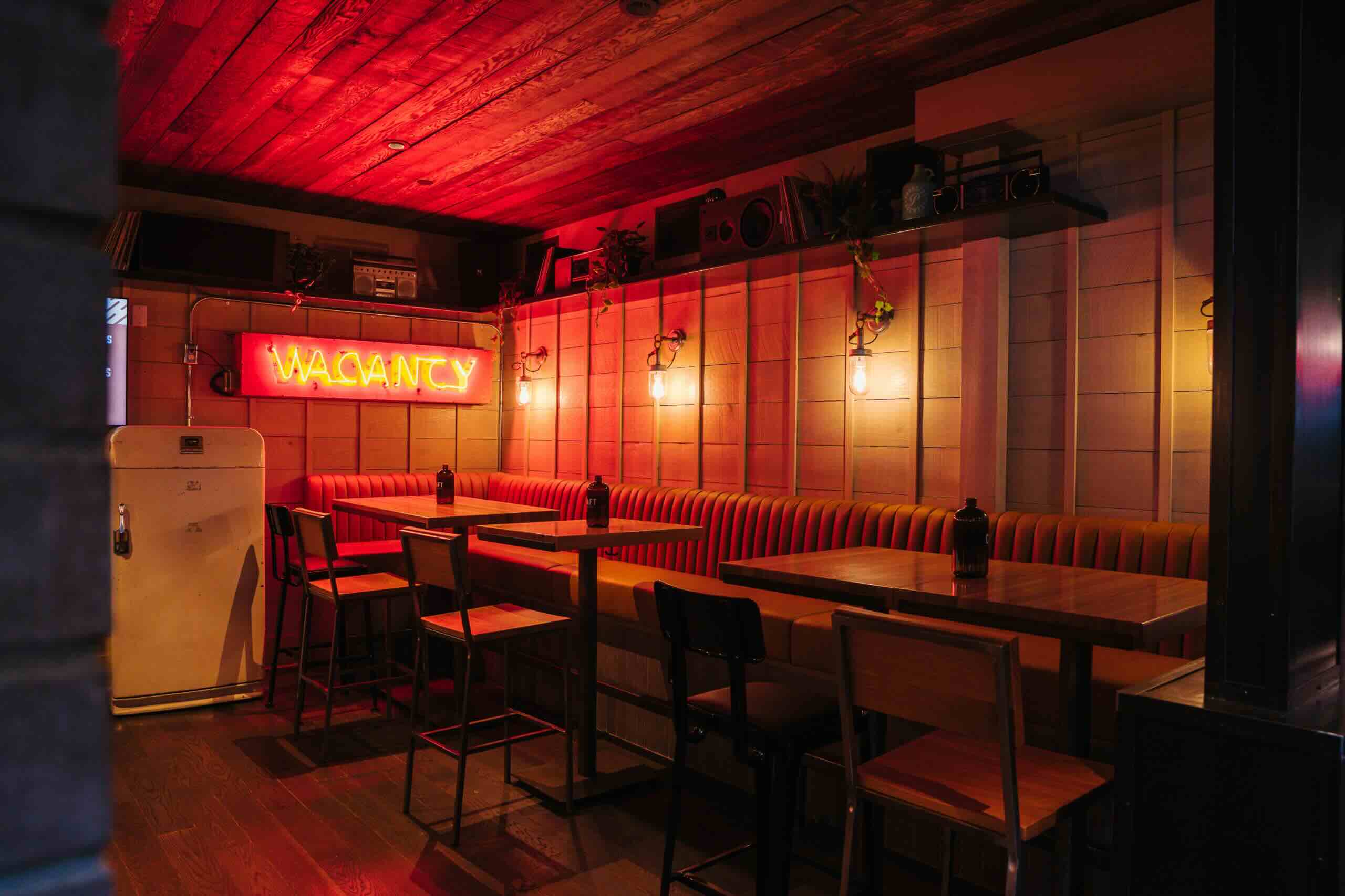CRAFT Beer Market Dimly lit restaurant interior with wooden tables, chairs, and booths. A neon sign reading "VACANCY" hangs on the wall above the seating area.