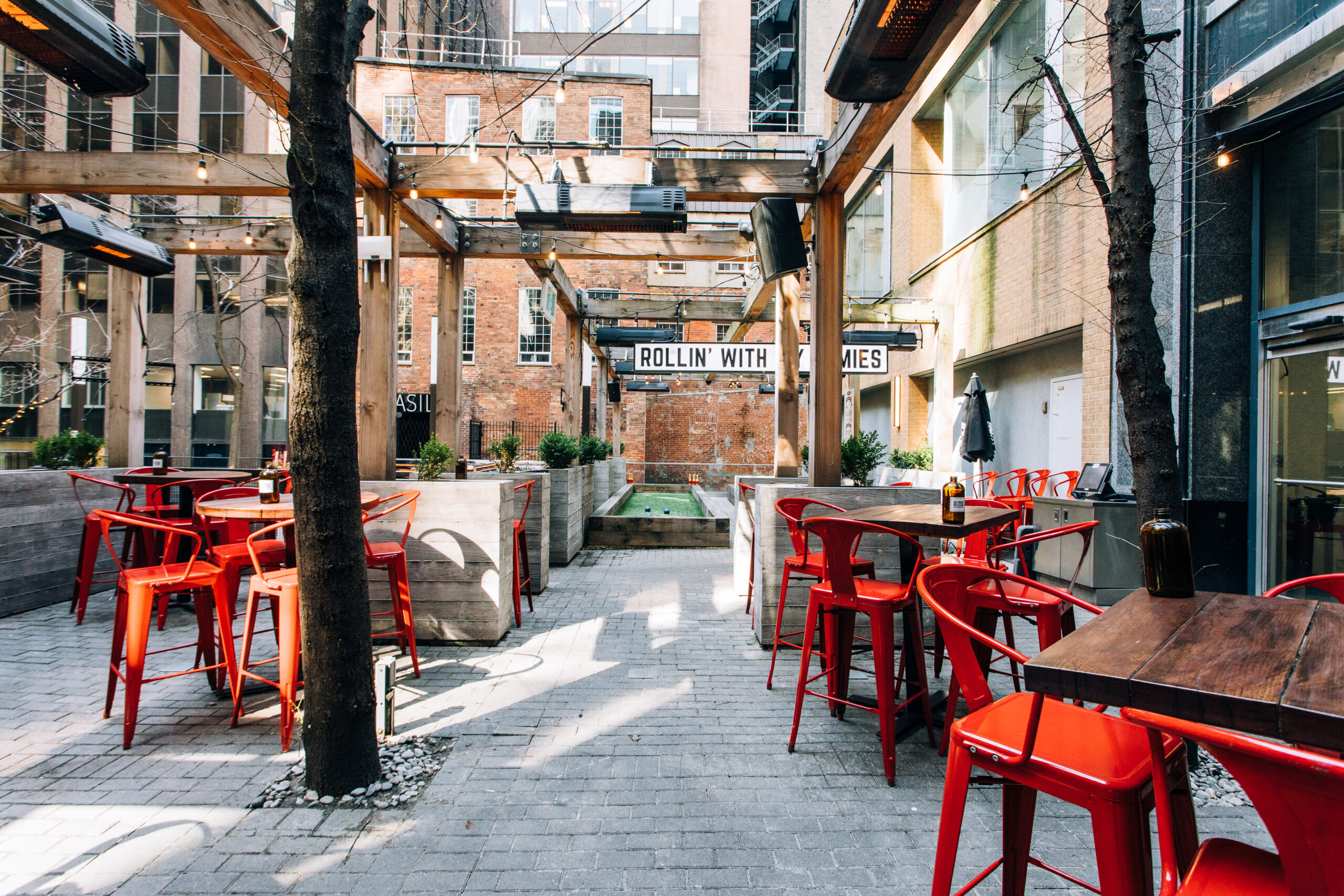 CRAFT Beer Market Outdoor patio with red chairs and wooden tables, surrounded by buildings; string lights hang above, a sign reads "ROLLIN' WITH MY HOMIES," and guests enjoy craft beer under the stars—perfect for private events.