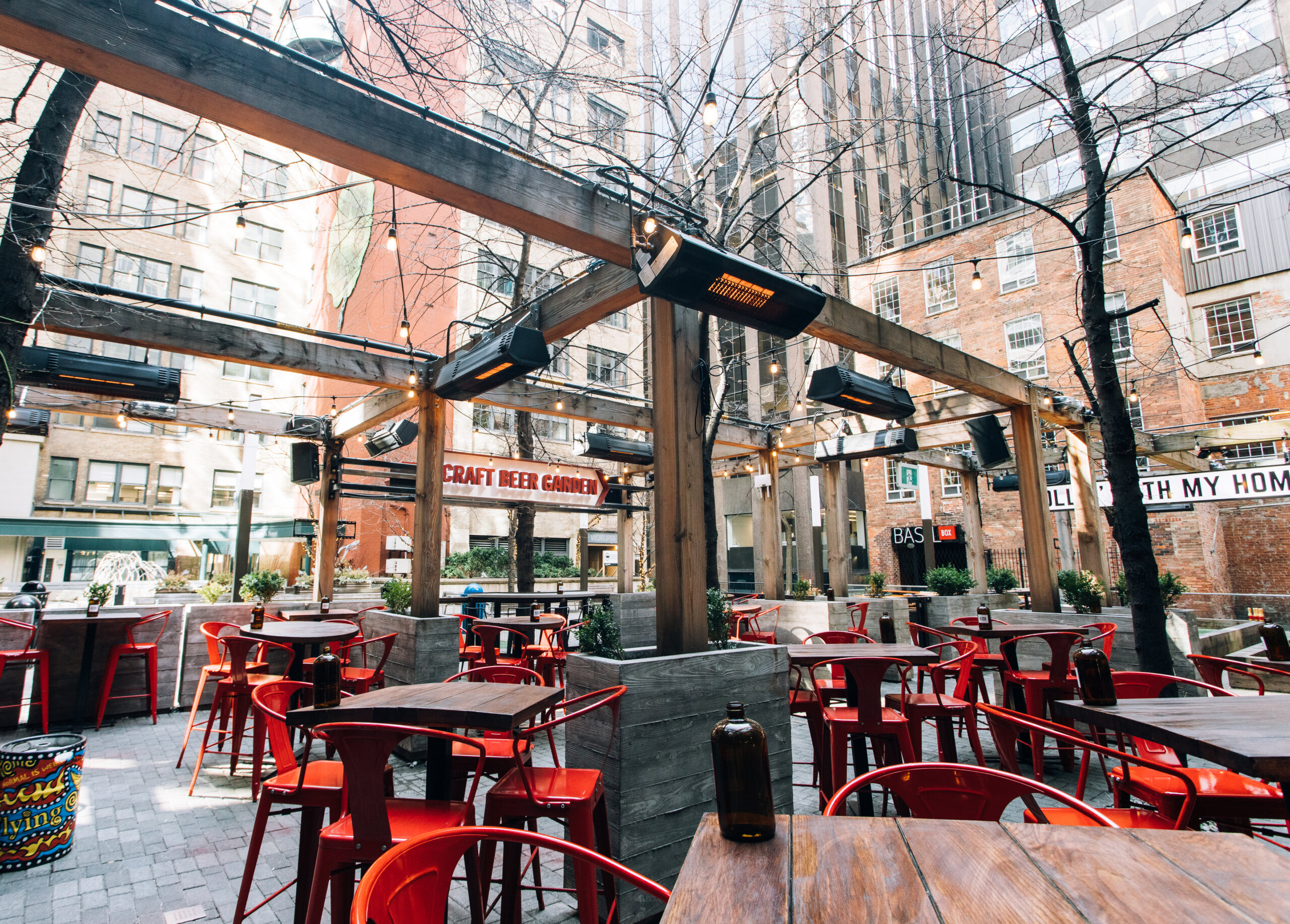 CRAFT Beer Market Outdoor urban beer garden with wooden tables and red chairs, surrounded by buildings, string lights overhead, heaters on wooden beams, and a menu featuring craft beer and cocktails. Perfect spot for group bookings.