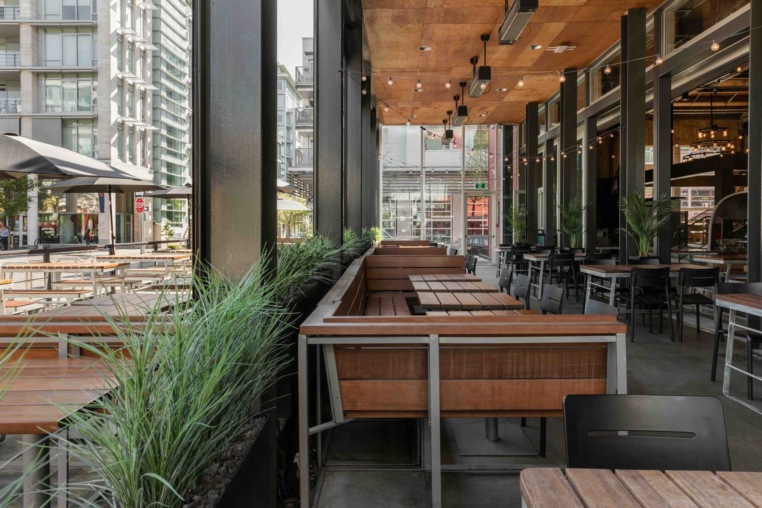 CRAFT Beer Market Modern outdoor and indoor dining area with wooden tables and chairs, surrounded by glass walls, plants, and city buildings in the background.