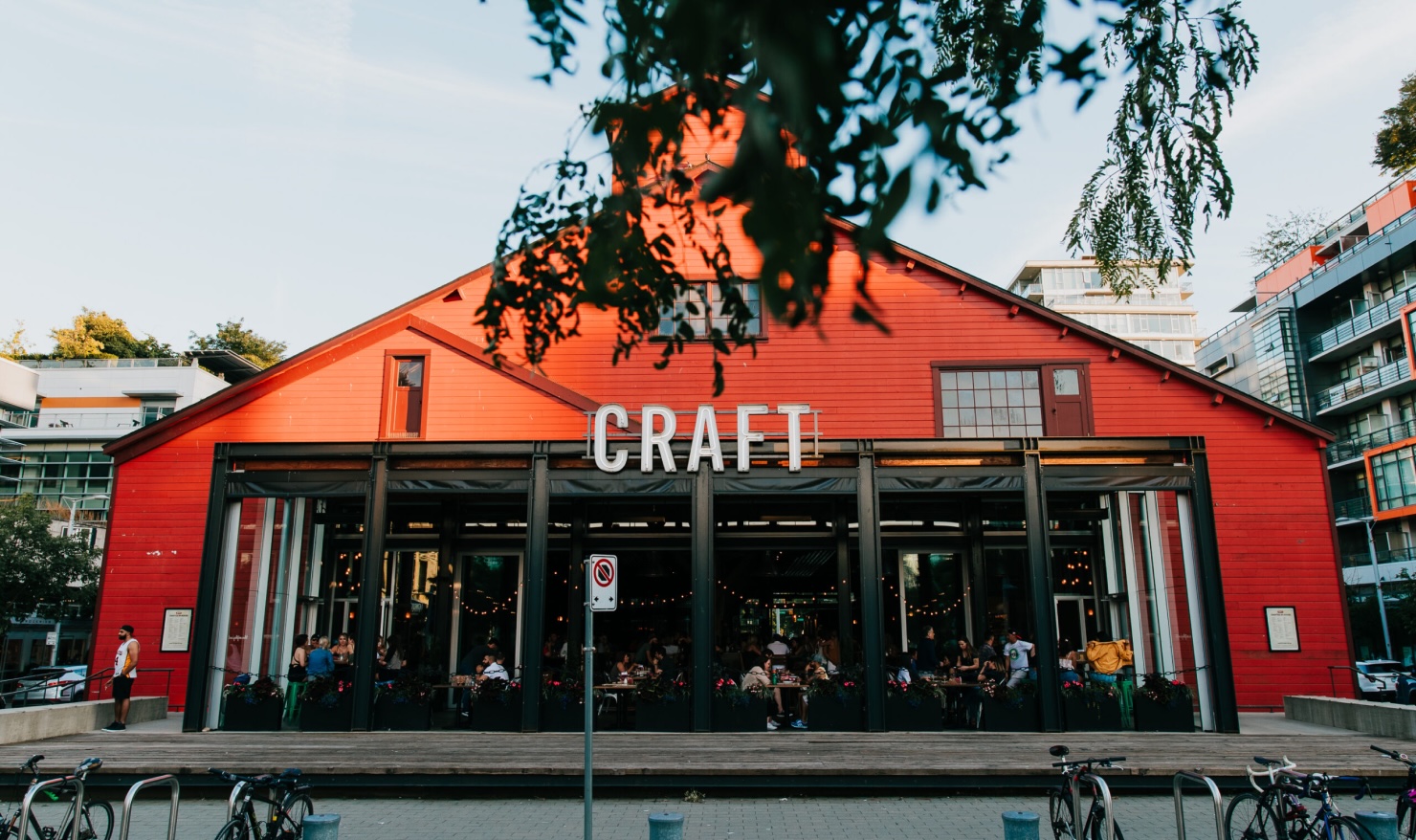 CRAFT Beer Market A red building with a peaked roof and large open windows has a sign reading "CRAFT" above the entrance; people are seated inside and bicycles are parked outside.