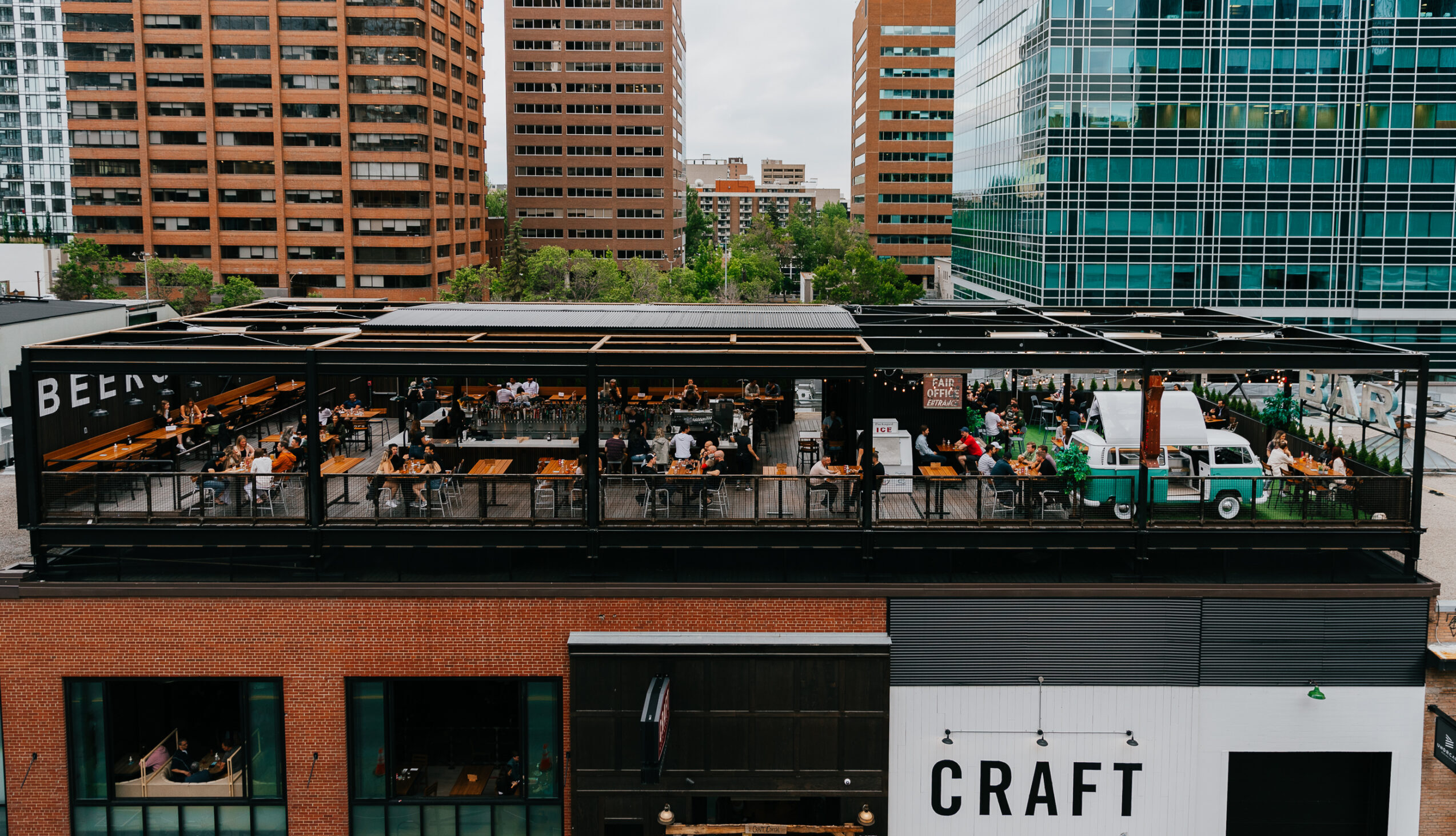 CRAFT Beer Market A rooftop restaurant with many people sitting at tables; a green and white truck is parked on the patio, surrounded by city buildings—perfect for group bookings and enjoying craft beers.