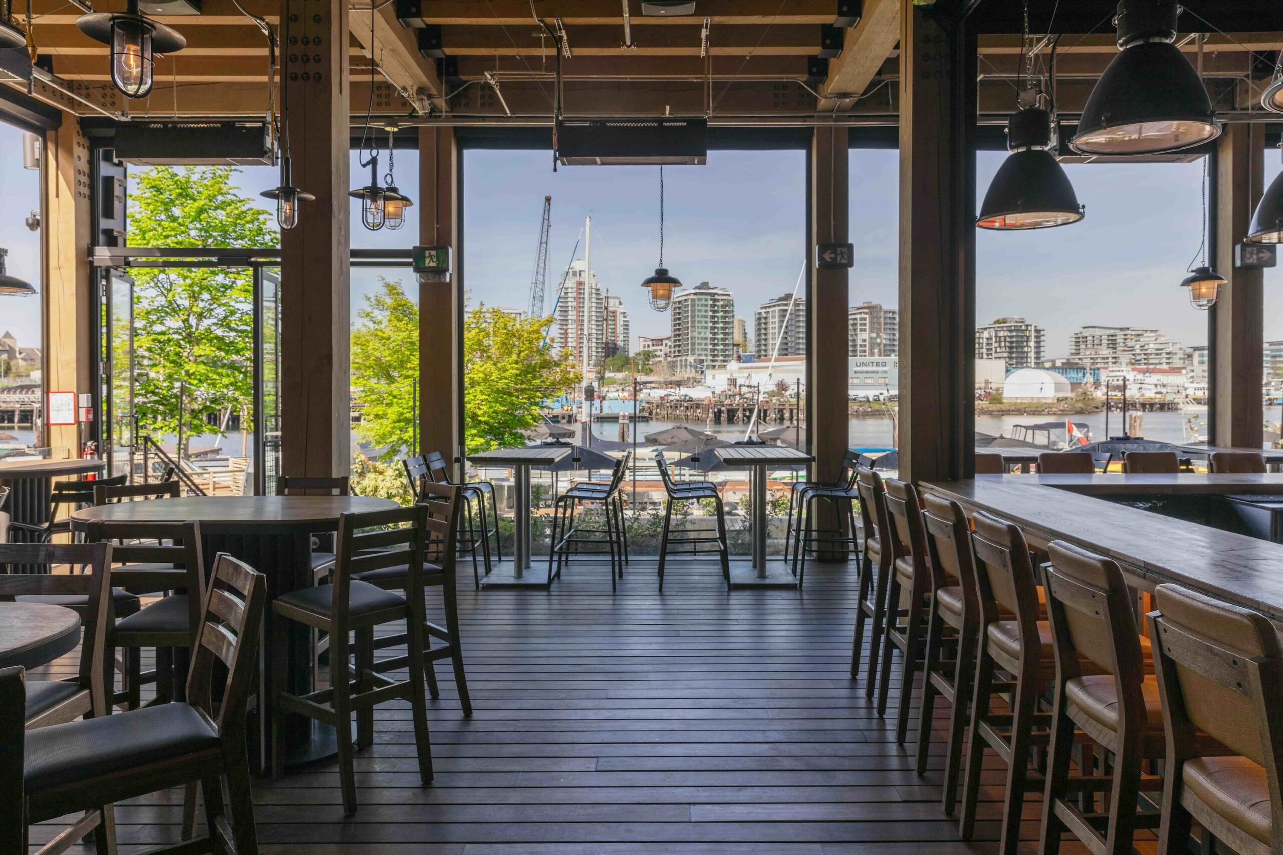 CRAFT Beer Market Modern restaurant interior with wooden floors and chairs, large windows overlooking a waterfront cityscape with high-rise buildings and a bridge, perfect for casual dining and vibrant social vibes.