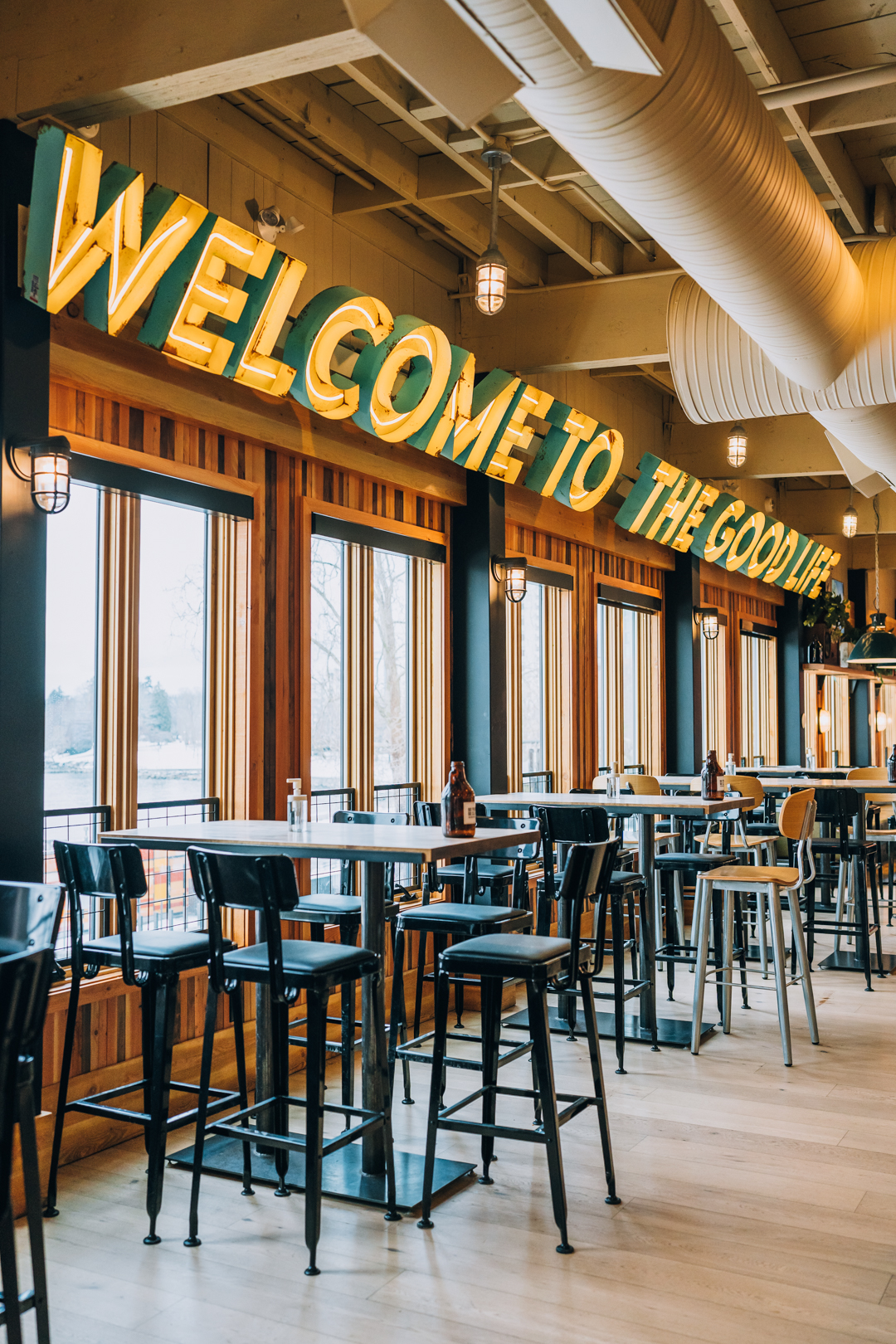 CRAFT Beer Market A restaurant interior with high tables and stools, large windows, and a lit sign reading "WELCOME TO THE GOOD LIFE" above the seating area, perfect for enjoying craft beer or hosting private events.