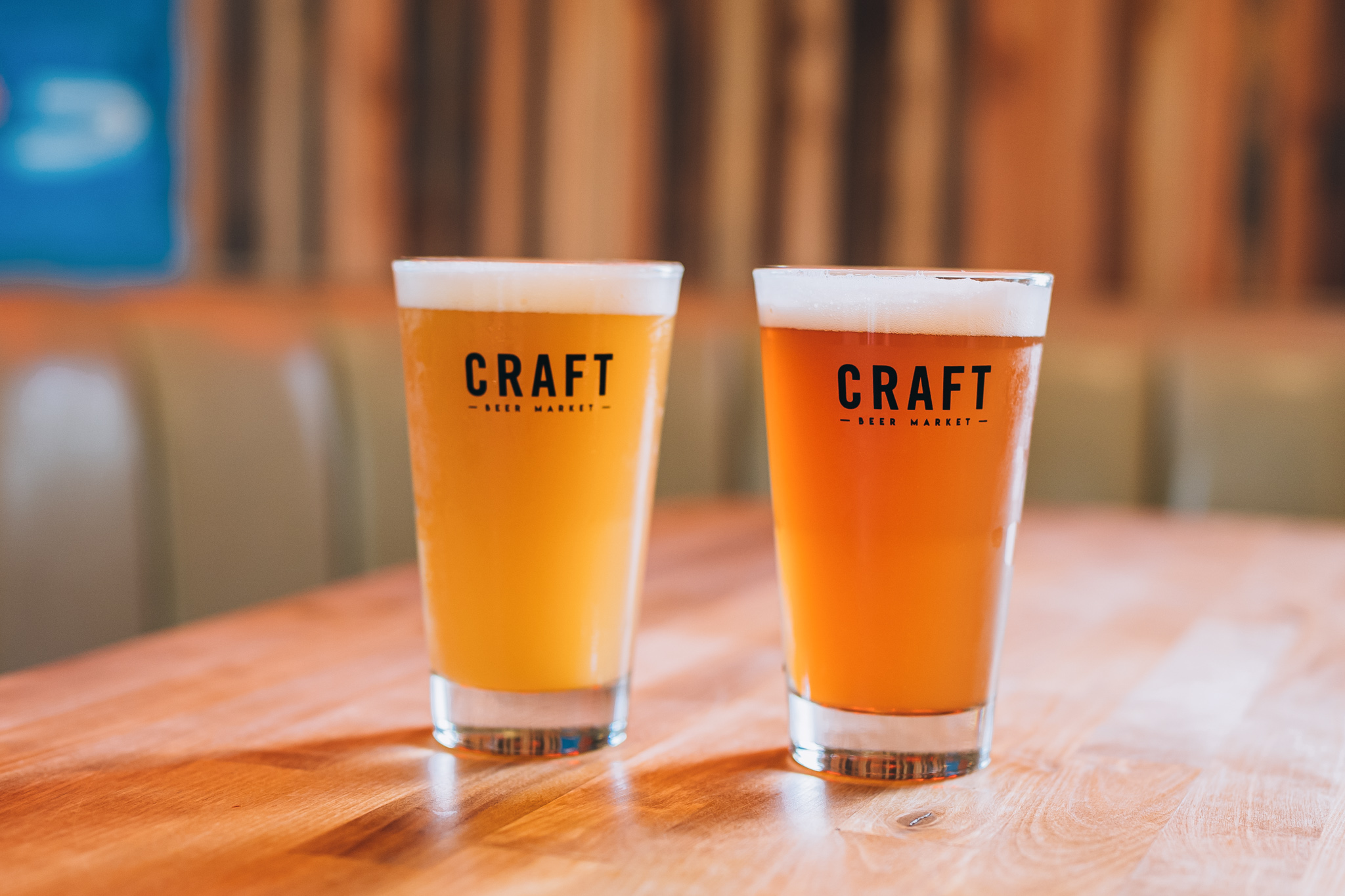 CRAFT Beer Market Two glasses of craft beer with frothy heads sit on a wooden table at this cozy beer garden, each glass labeled "CRAFT Beer Market." The background is softly blurred.