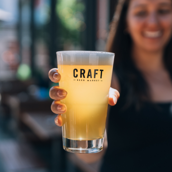 CRAFT Beer Market A person holds out a pint glass of hazy craft beer with "CRAFT BEER MARKET" printed on it; background is slightly blurred. Perfect for group bookings or enjoying unique craft beers with friends.