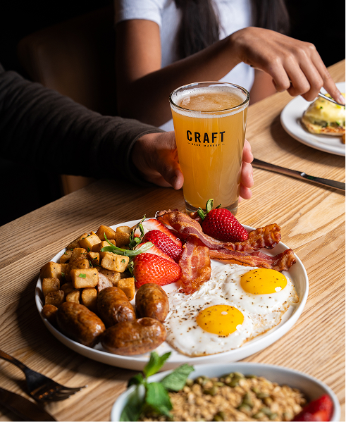 CRAFT Beer Market A plate with eggs, bacon, sausage, potatoes, and strawberries sits on a wooden table. A person holds a glass of beer labeled "CRAFT" beside the plate—perfect for group bookings at one of our multiple locations.