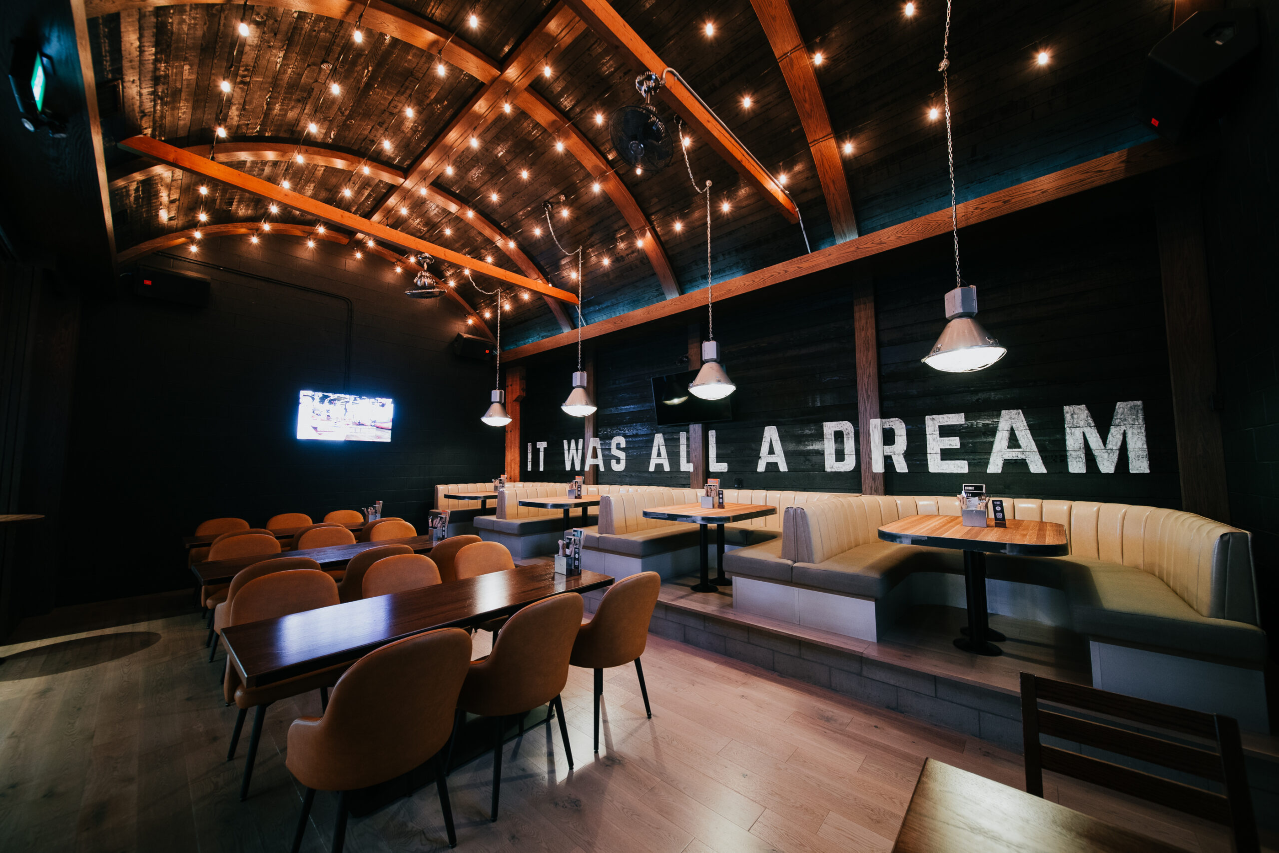 CRAFT Beer Market A modern restaurant interior with wooden tables, yellow chairs, booth seating, and string lights. Enjoy craft cocktails beneath a sign that reads “IT WAS ALL A DREAM.” Perfect for group bookings.