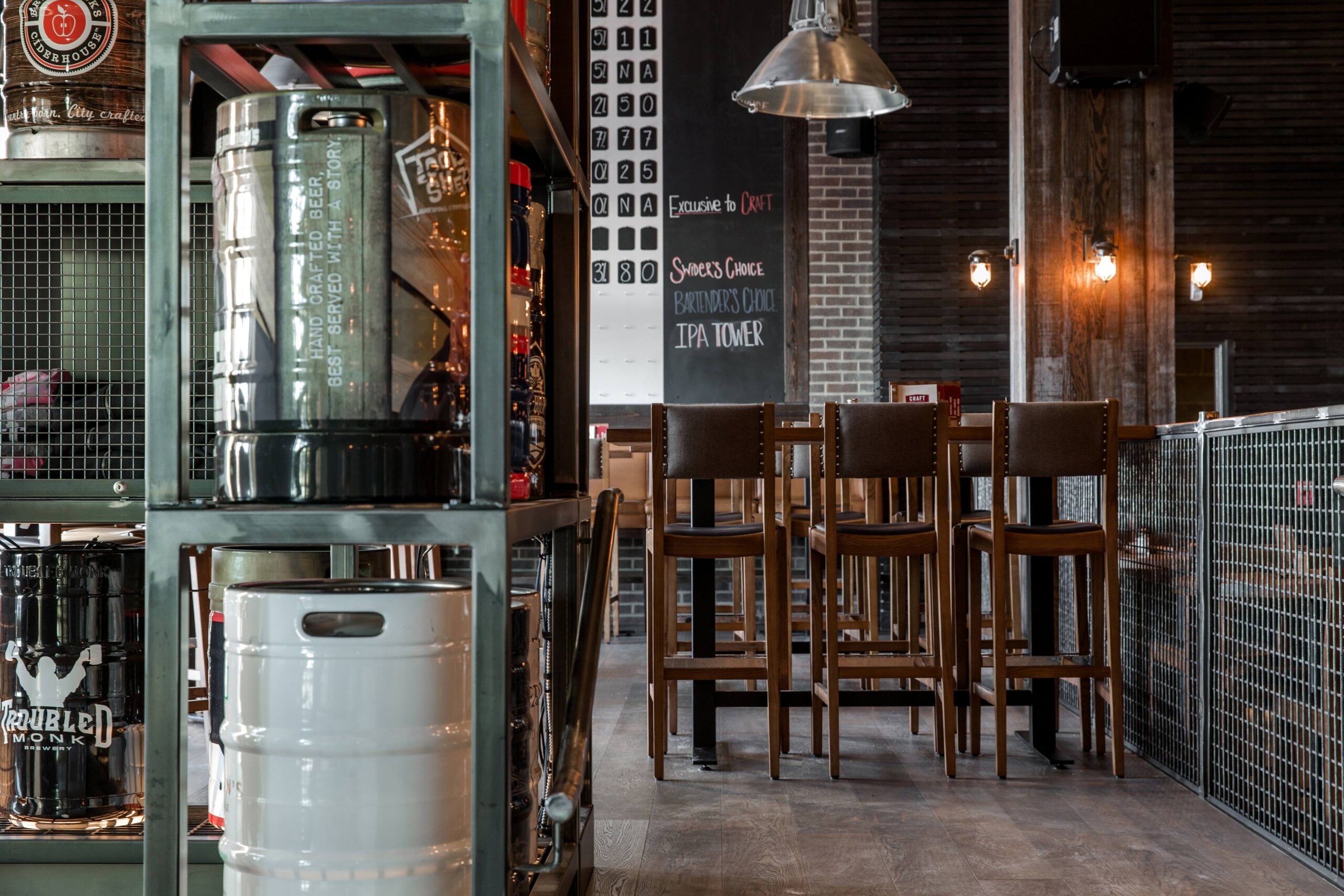 CRAFT Beer Market Metal racks with beer kegs, high wooden chairs, and a chalkboard menu in a modern industrial-style bar or brewery interior.