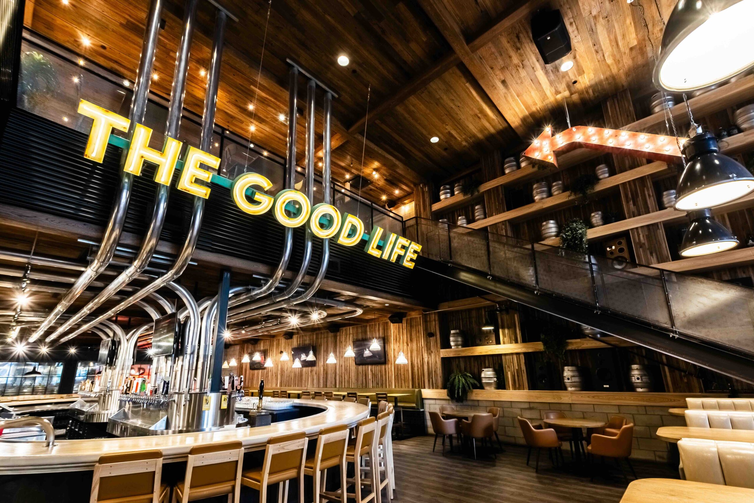 CRAFT Beer Market A modern bar with wood-paneled walls, curved counter seating, and a large neon sign reading "THE GOOD LIFE" above the bar area.