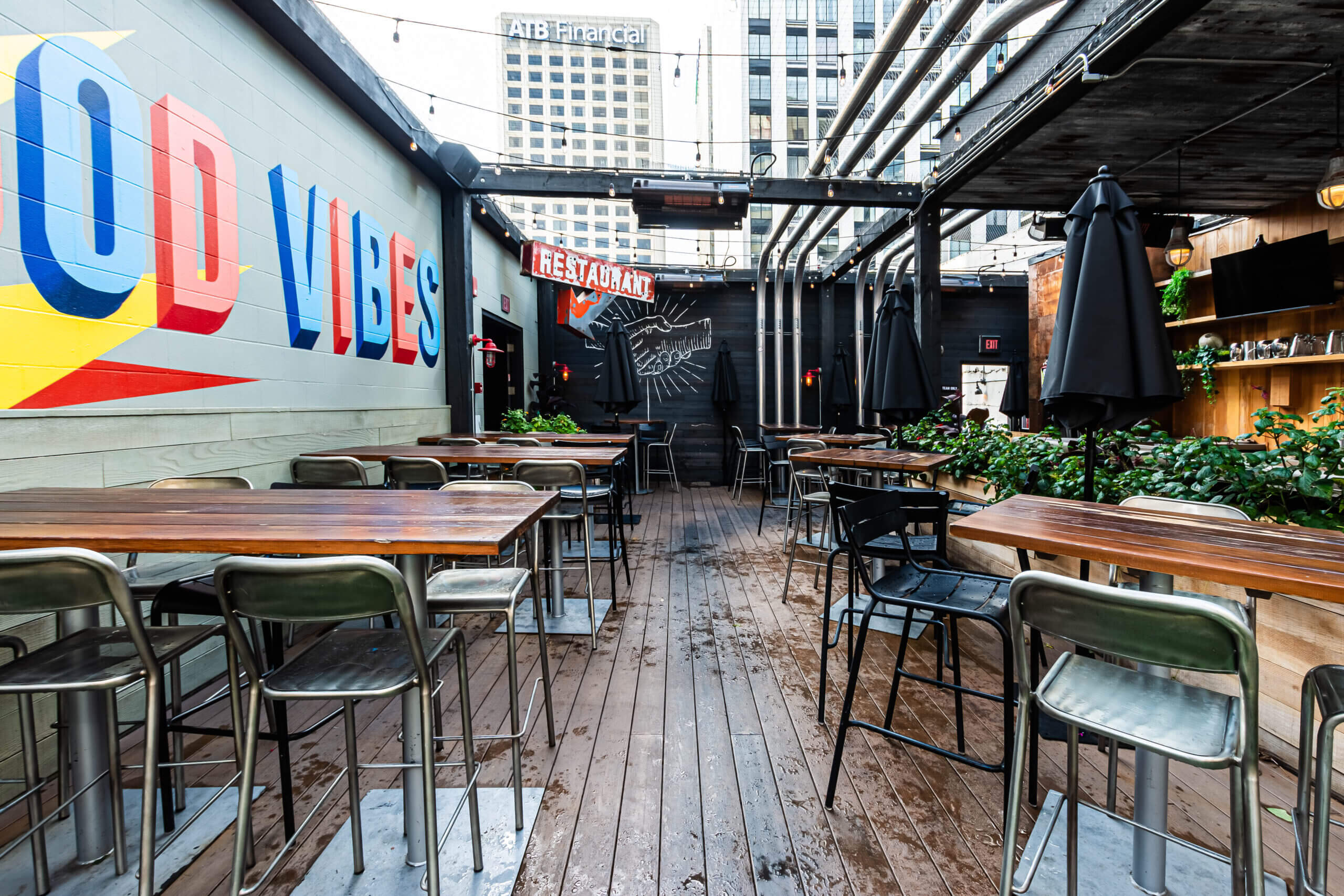 CRAFT Beer Market Outdoor restaurant patio with wooden tables, metal chairs, string lights, potted plants, and wall art reading "GOOD VIBES" in an urban setting—perfect for group bookings and enjoying craft beers or cocktails.