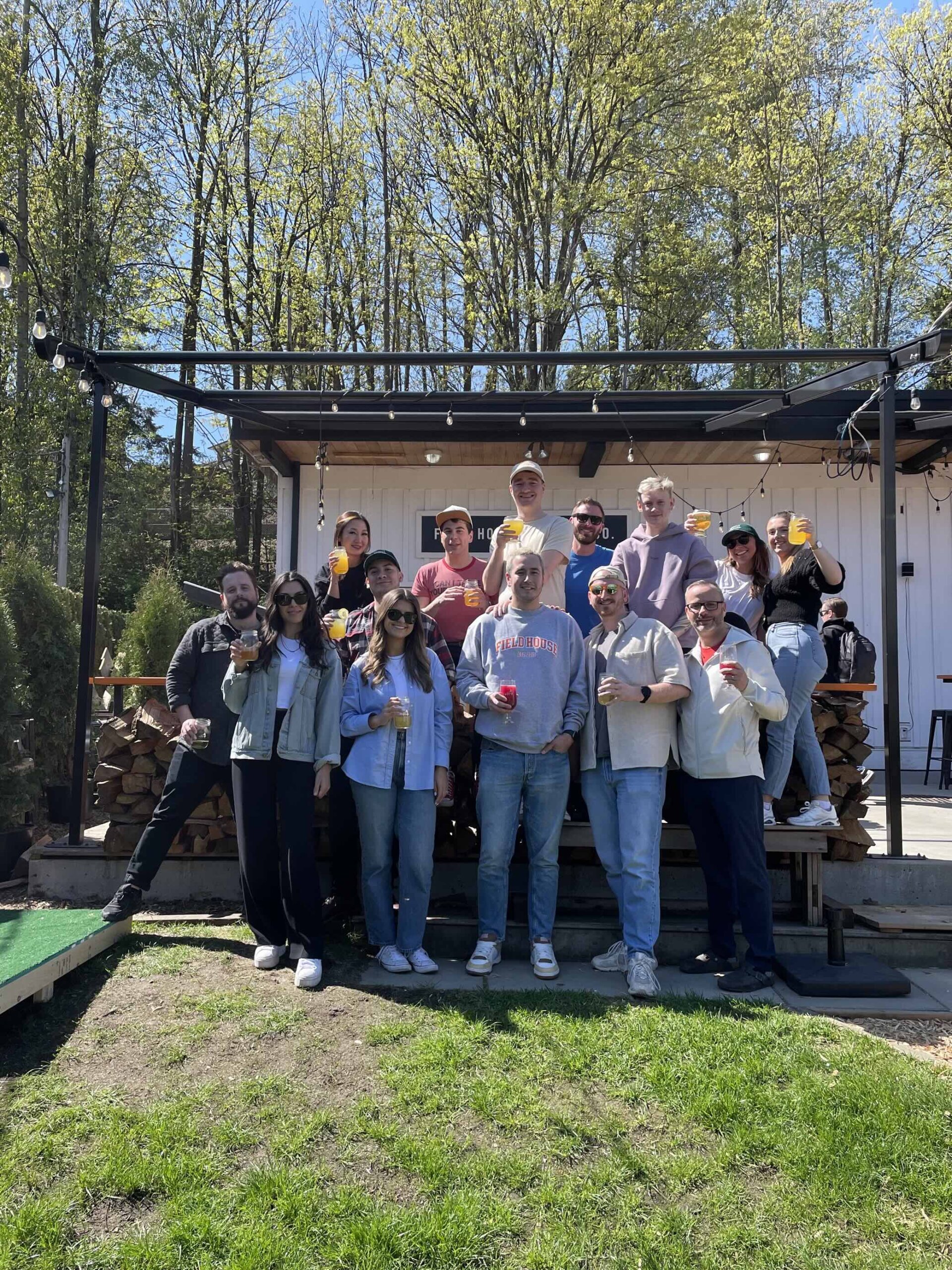 CRAFT Beer Market A group of fifteen people stands outdoors in front of a white building, holding drinks and posing for a photo on a sunny day. Trees and stacked firewood are visible in the background.