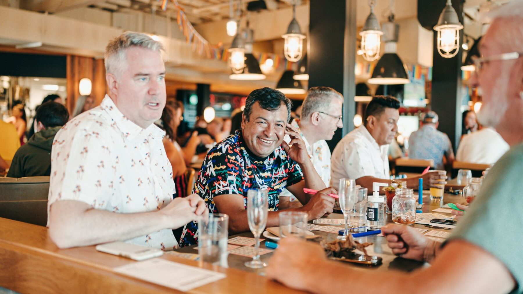 CRAFT Beer Market A group of people sit at a bar table, talking and smiling, with craft cocktails, food, and bingo cards in front of them in a lively indoor setting—perfect for group bookings.