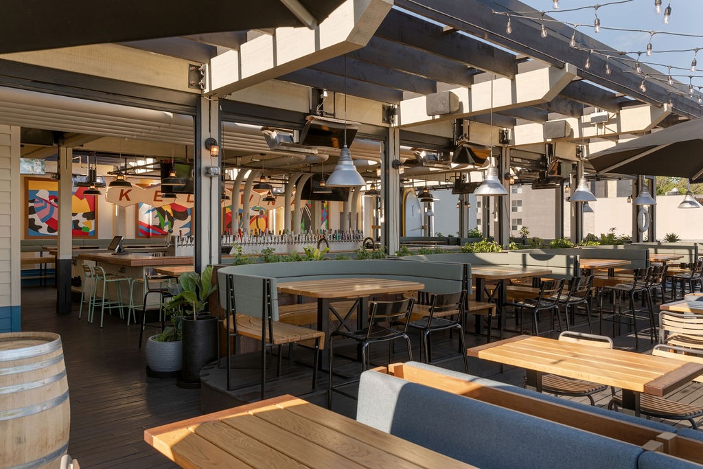 CRAFT Beer Market Outdoor restaurant patio with wooden tables, benches, and chairs under a pergola; string lights hang above and colorful wall art is visible in the background.