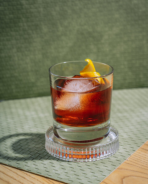 CRAFT Beer Market A glass of dark cocktail with a large ice cube and a lemon twist garnish sits on a glass coaster atop a patterned surface, perfect for soaking in social vibes during casual dining.