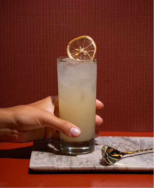 CRAFT Beer Market A hand holds a tall glass of a light-colored cocktail with ice and a dried lemon wheel garnish, placed on a marble slab next to a gold cocktail spoon—perfect for group bookings at our multiple locations.