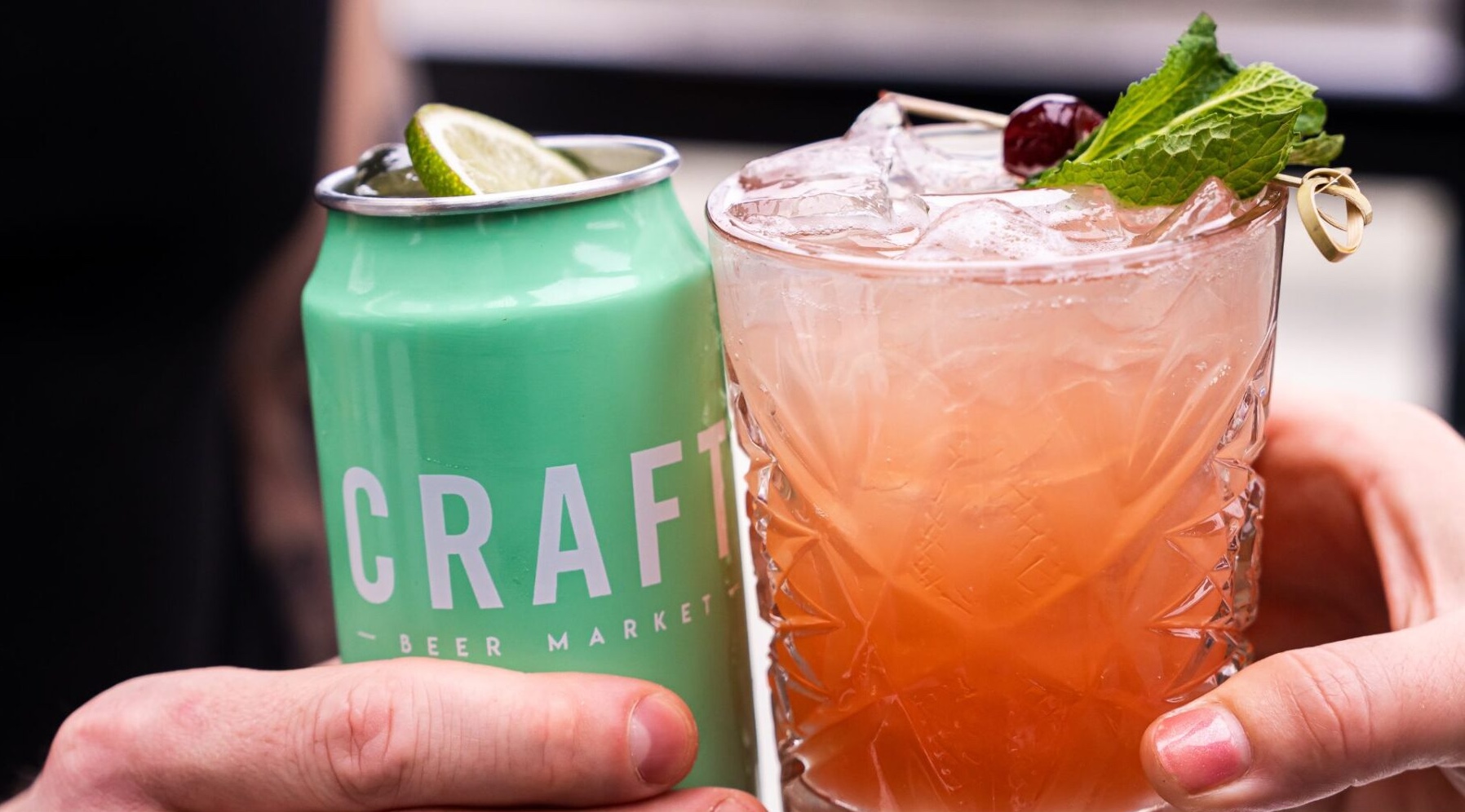 CRAFT Beer Market A hand holds a green can labeled "CRAFT" next to another hand holding a cocktail with ice, a cherry, mint, and a lime garnish—perfect for enjoying craft cocktails with friends or group bookings.