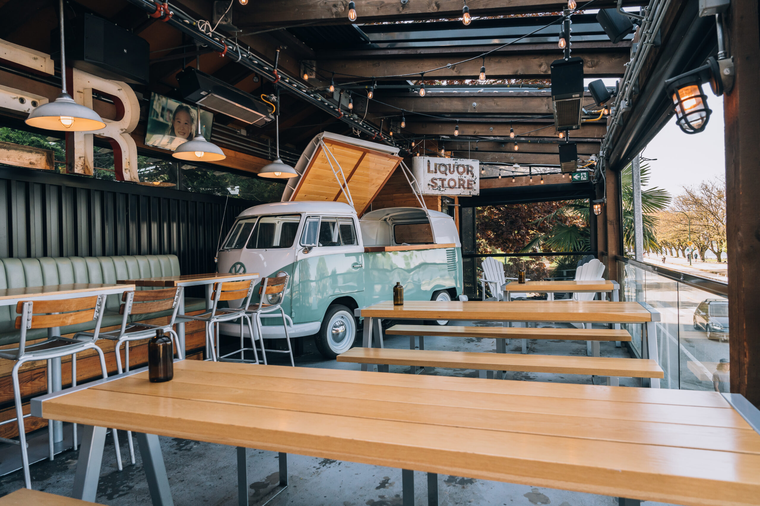 CRAFT Beer Market Outdoor seating area of a cafe with wooden tables, benches, tall stools, and a retro van converted into a bar serving craft beers, under a covered patio with string lights. Perfect for group bookings.