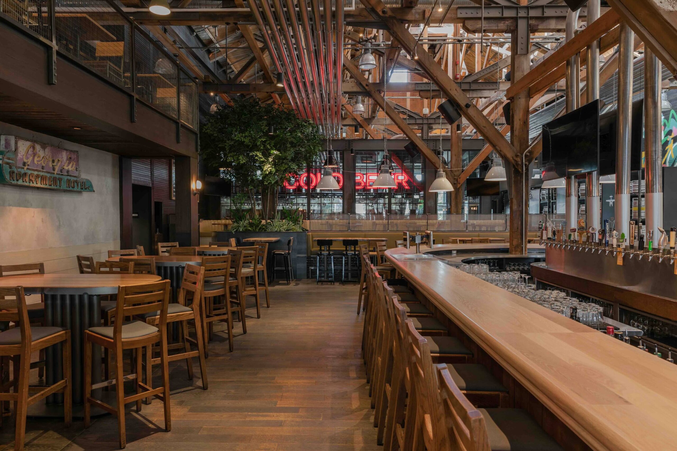 CRAFT Beer Market Spacious bar interior with wooden tables and chairs, a long bar counter, taps, indoor plants, and large windows letting in natural light—perfect for casual dining and enjoying social vibes.