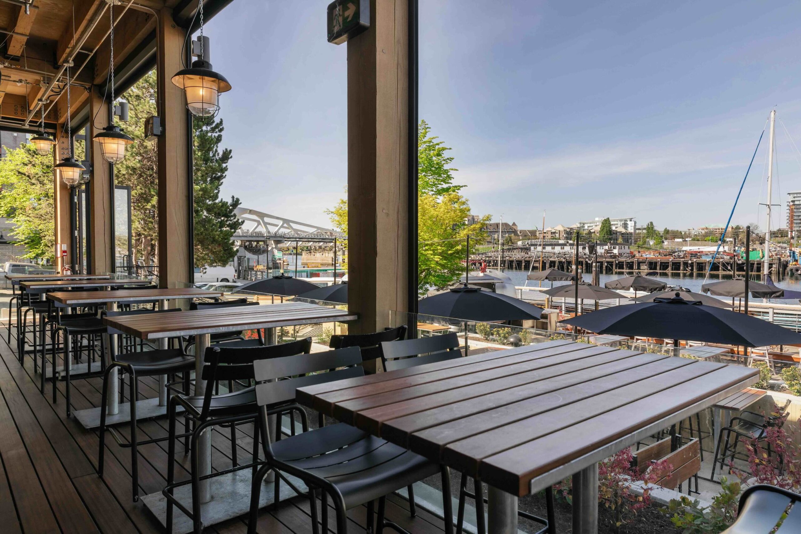 CRAFT Beer Market Open-air restaurant patio with wooden tables and metal chairs overlooking a waterfront area, perfect for casual dining while enjoying craft cocktails under umbrellas, with boats and a bridge in the background.