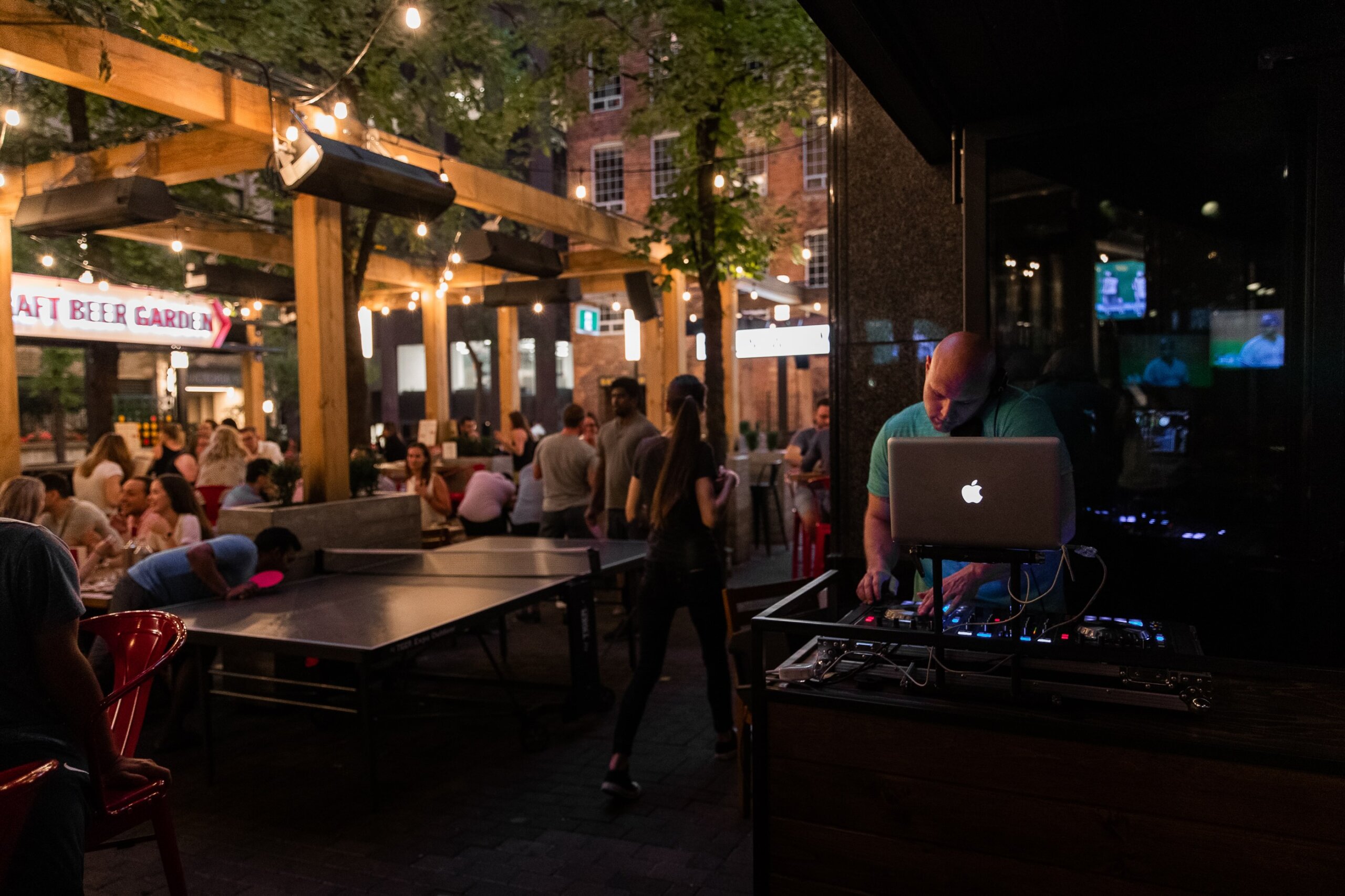 CRAFT Beer Market A DJ plays music on a laptop while people sit and socialize at outdoor tables under string lights in a beer garden at night.
