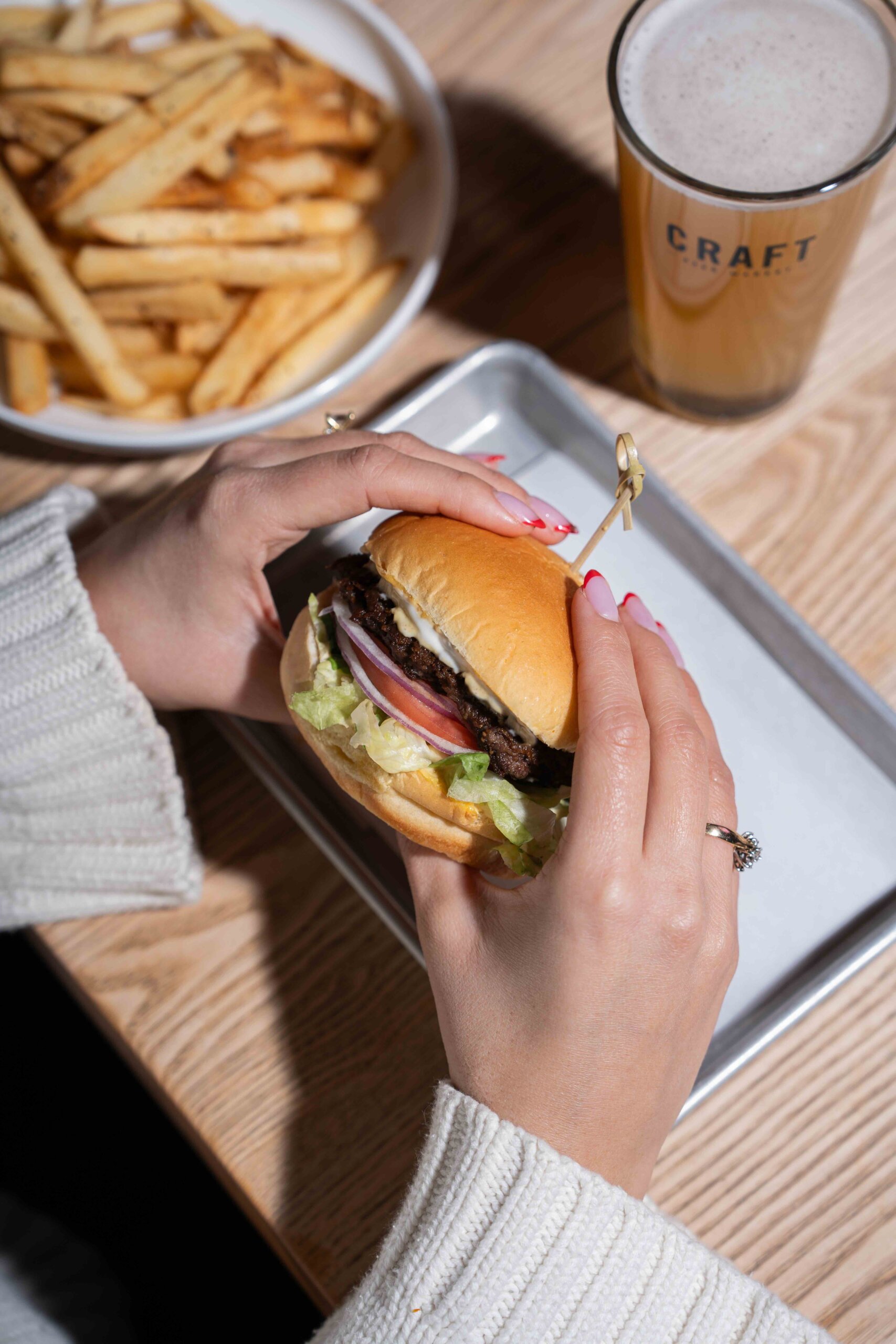 CRAFT Beer Market Person holding a hamburger with lettuce, tomato, and onion, next to a tray, with a bowl of fries and a glass of beer on a wooden table.