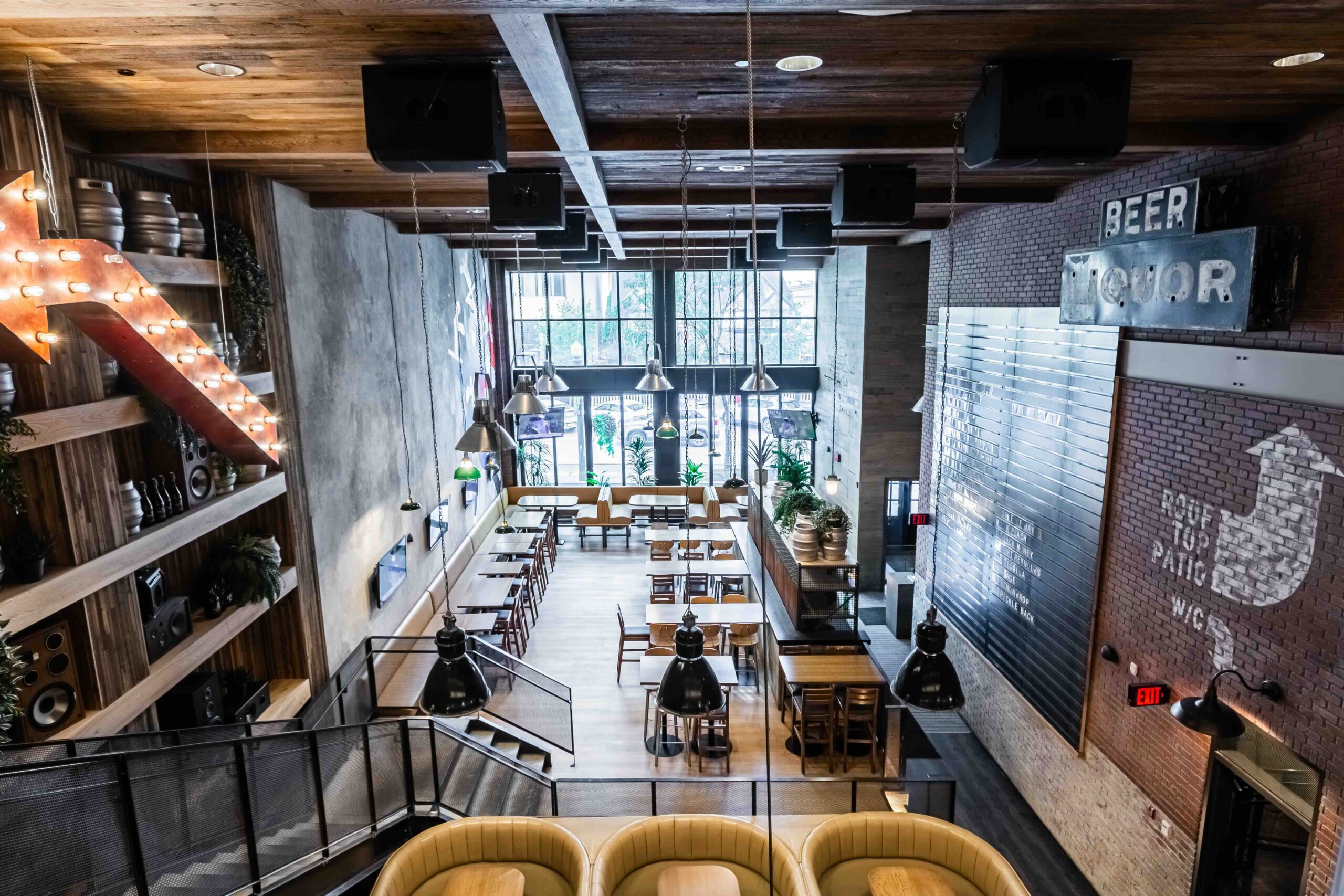 CRAFT Beer Market Modern restaurant interior with large windows, wooden tables and chairs, industrial lighting, and decorative signs on the walls. View is from an upper level overlooking the dining area.